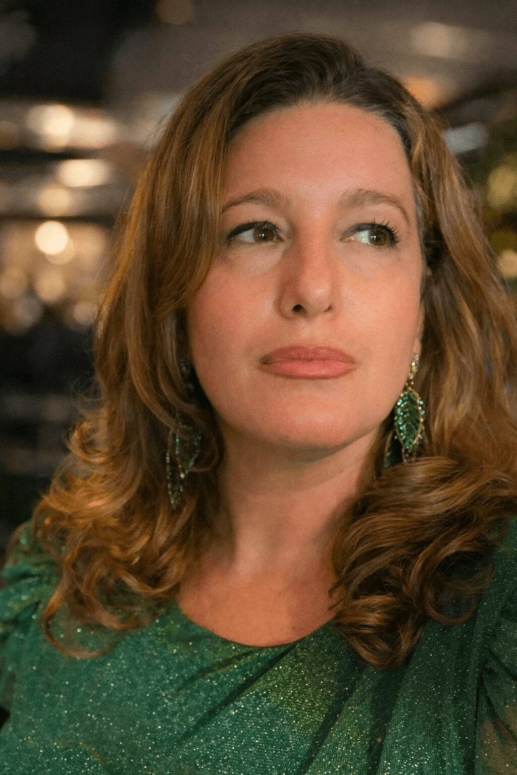 Woman with wavy hair wearing green earrings and a green top, looking to the side.