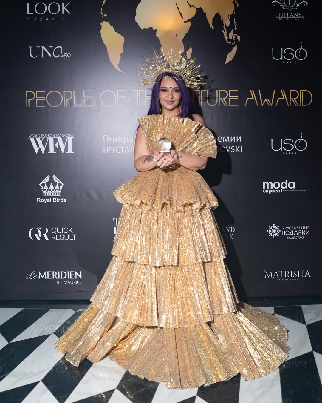Woman in a gold, layered, floor-length gown holding an award, standing on a black and white checkered floor.