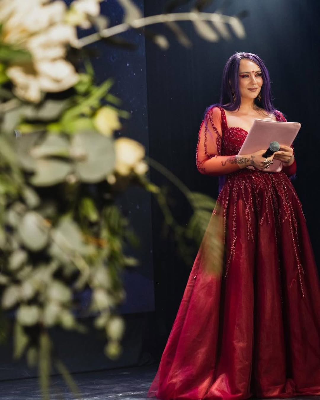 Woman in a red gown holding a tablet on stage with floral decorations in foreground.