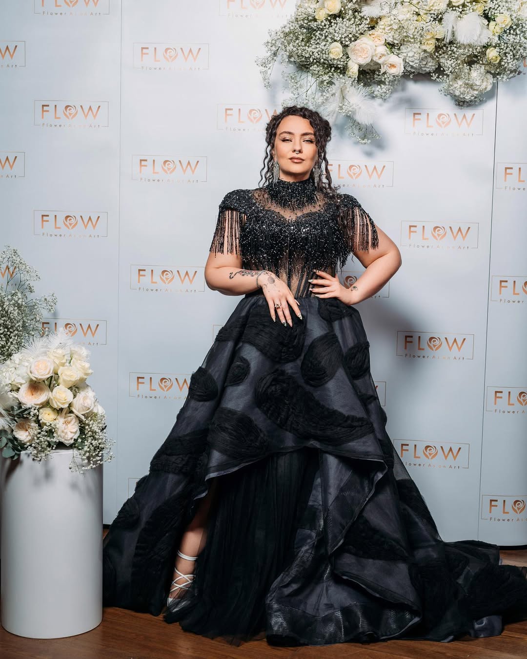 Woman in a black gown with lace top and full skirt, standing with hands on hips, in front of a floral backdrop.