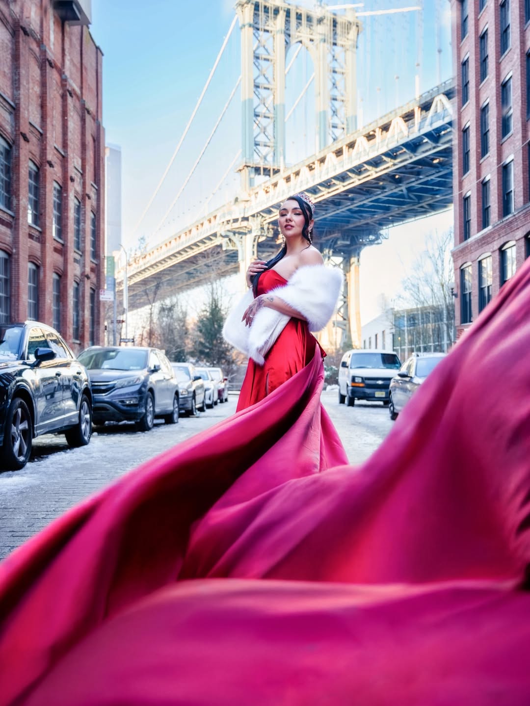 Woman in red gown standing on city street with bridge overhead and cars parked nearby.