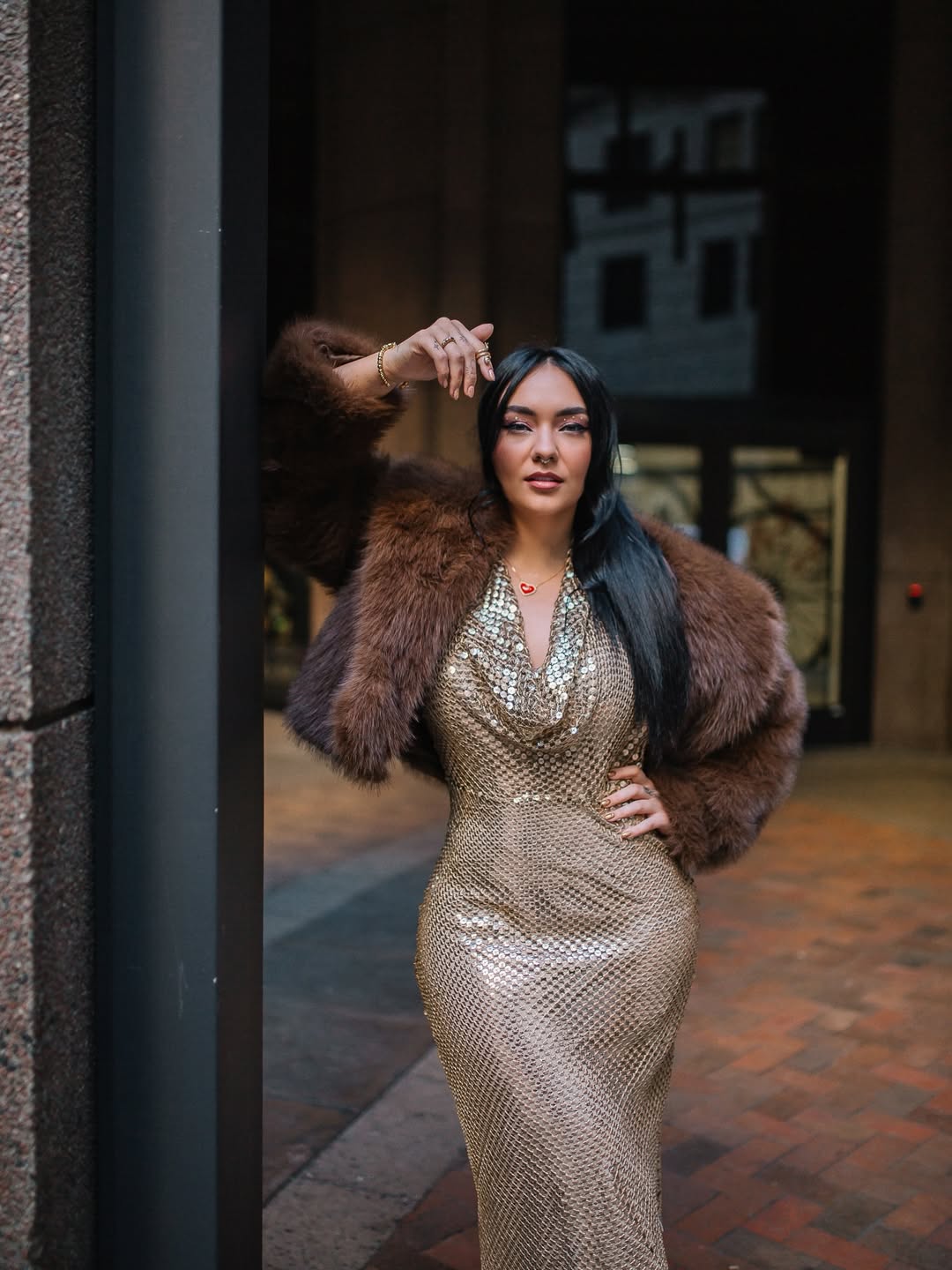 Woman in a shiny, patterned dress and fur coat standing outdoors, with a building in the background.