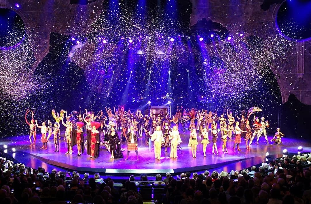 Large stage with performers, colorful lights, and confetti, audience watching a lively show.