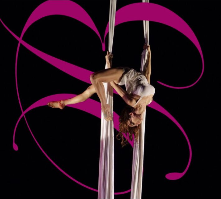 A person performs an aerial silk routine, hanging upside down with legs extended and arms gripping the fabric.