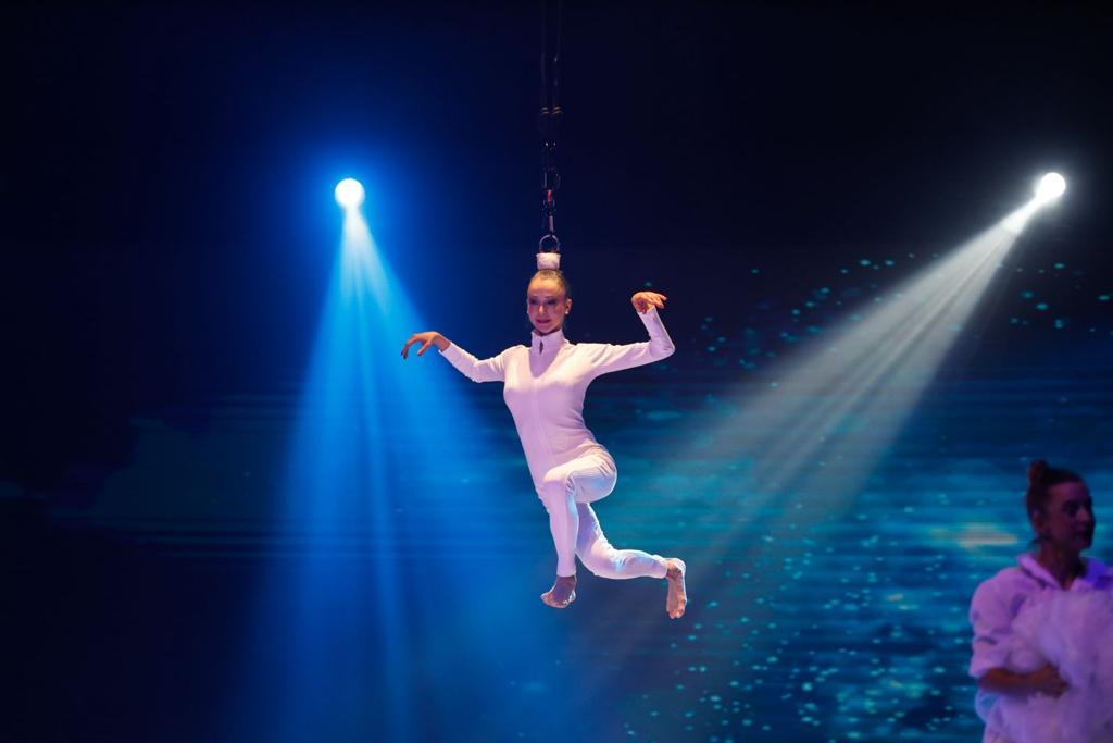 Performer in white costume hanging from a chain with arms raised, stage lights shining down, dark background.