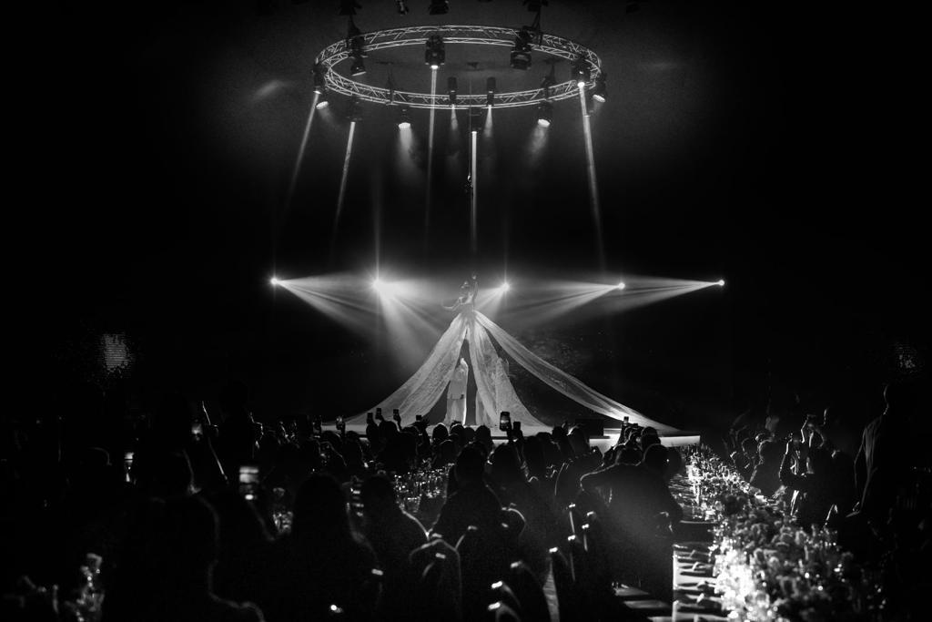 Crowd gathered at event with stage, circular lighting fixture, and draped fabric backdrop, illuminated by spotlights and stage lights.