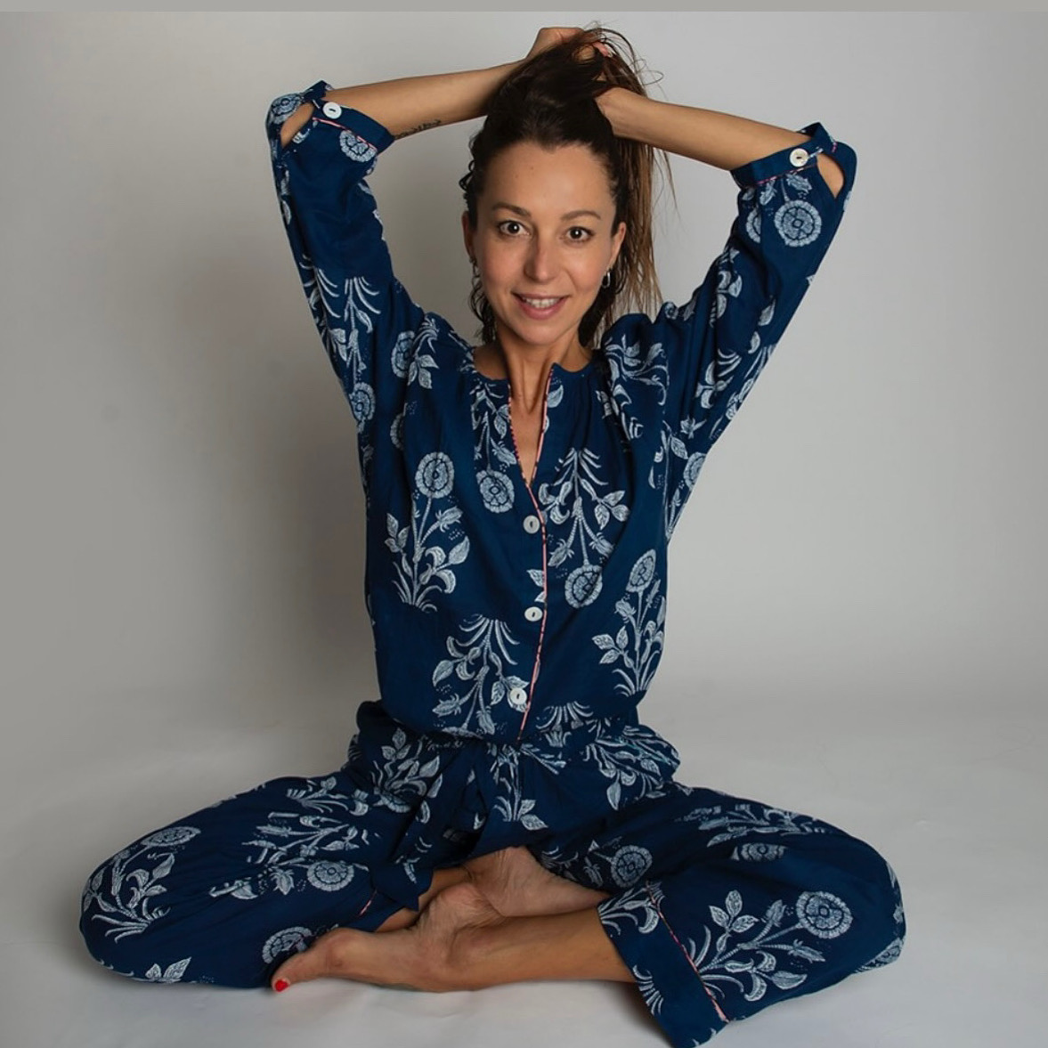 Woman sitting cross-legged on the floor, smiling, with hands in hair, wearing blue patterned pajamas, against a plain background.