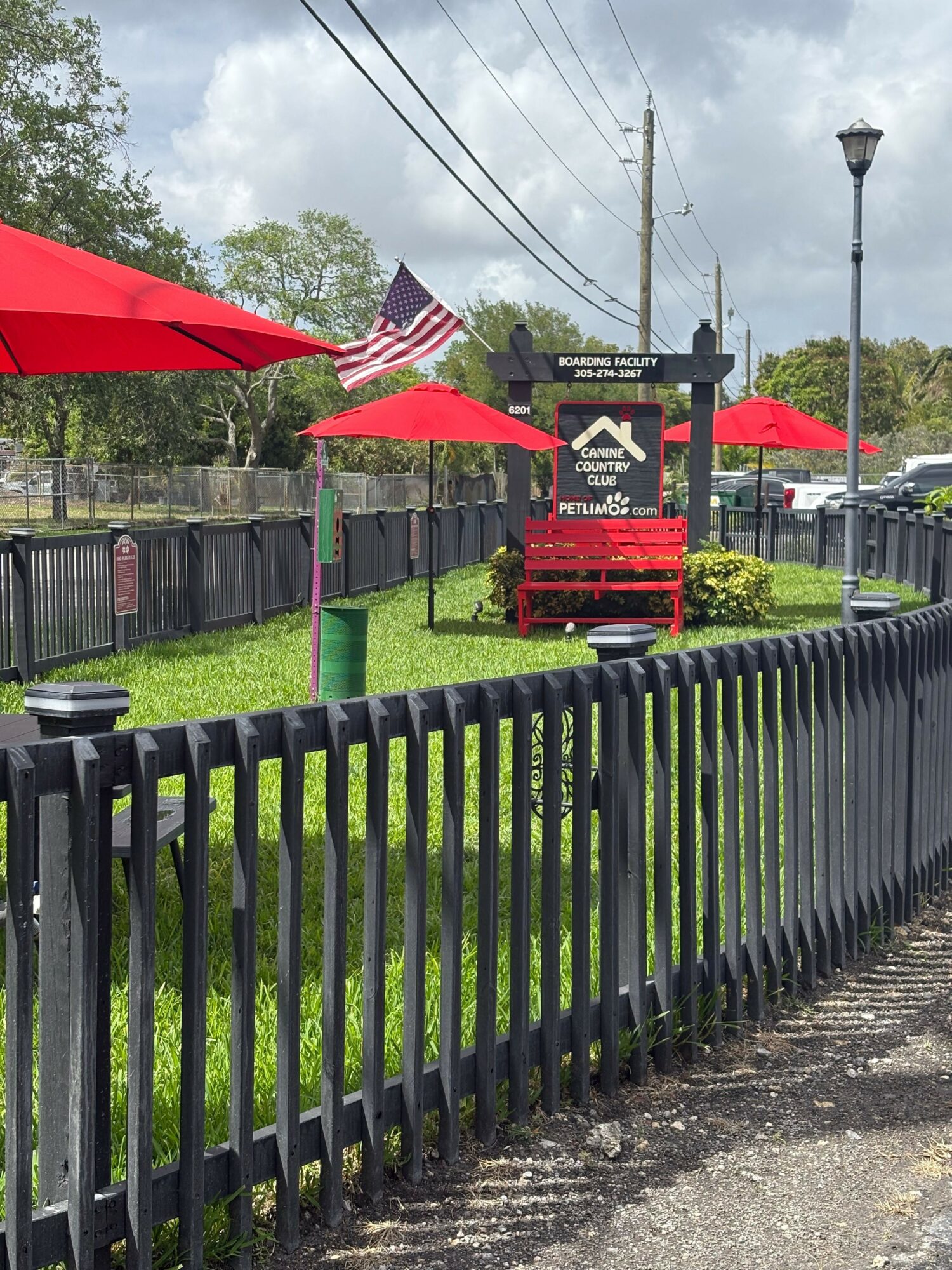 Outdoor seating area with red umbrellas, a black fence, and a sign in the background, with trees and a cloudy sky.