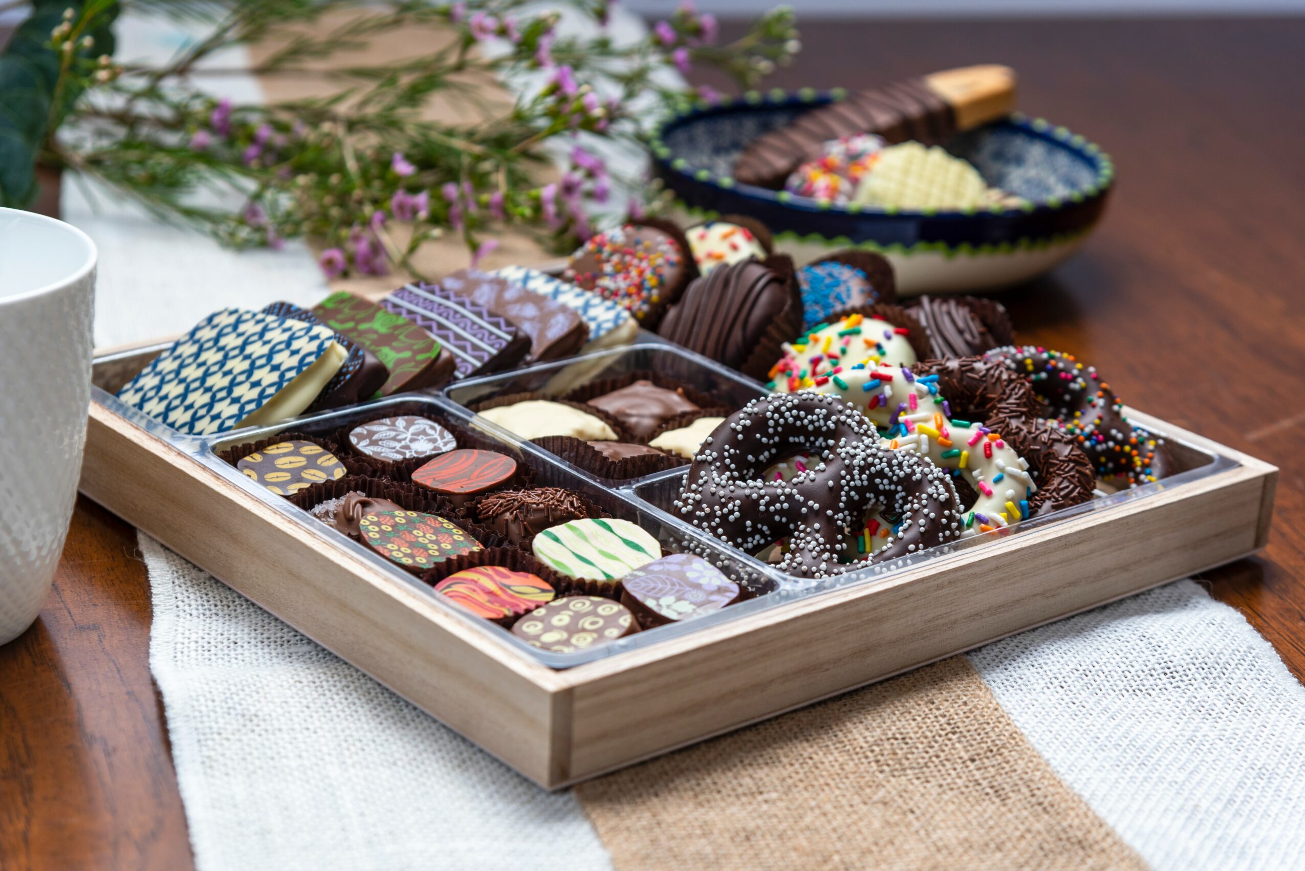 Tray of assorted chocolates and candies on a table with a bowl of candies and flowers in background.