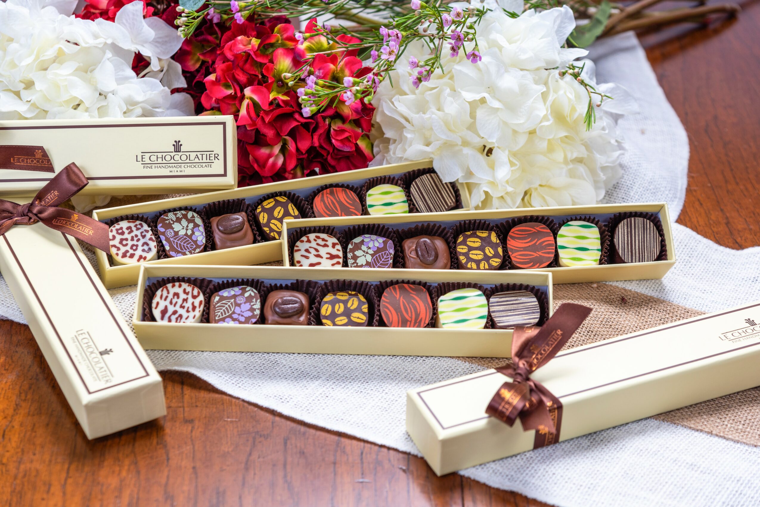 Open boxes of assorted chocolates on a wooden table with flowers in the background.