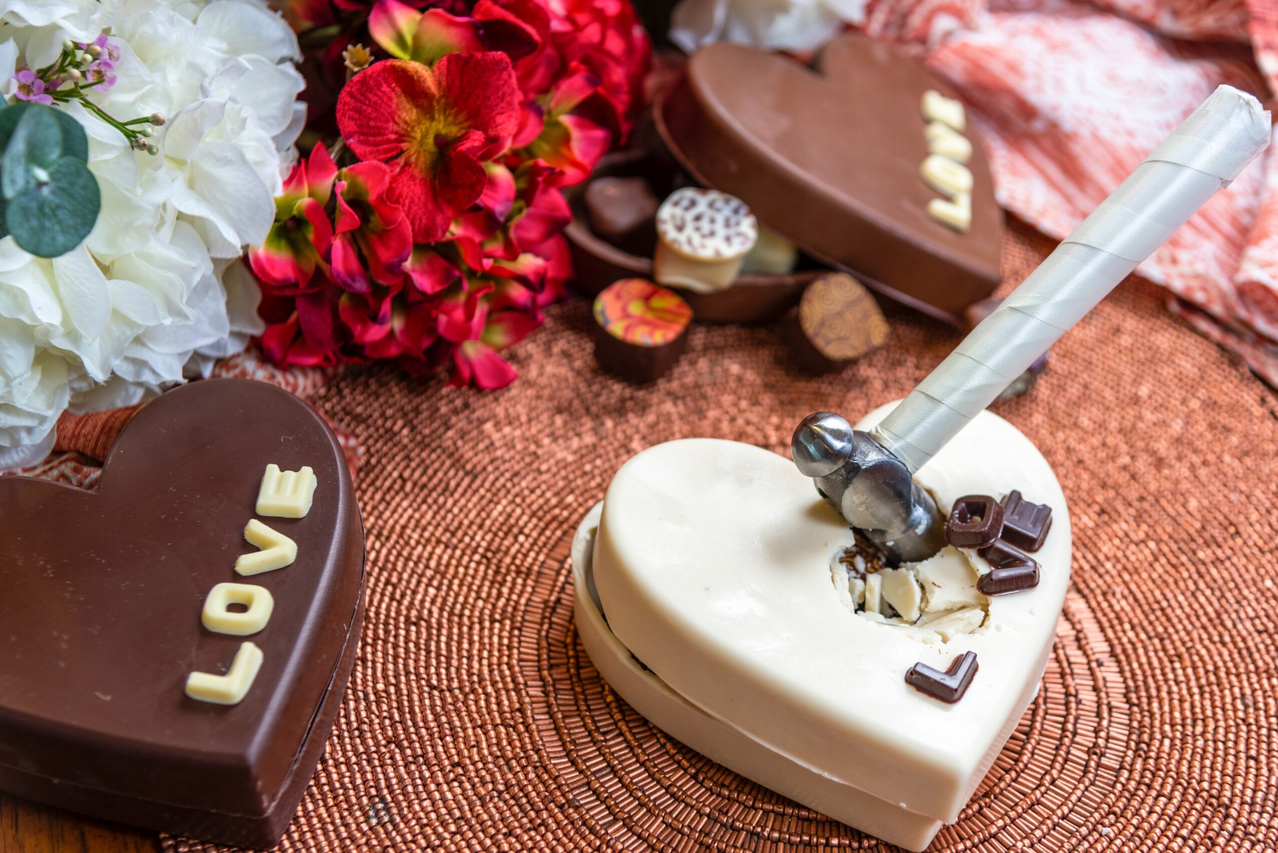 Heart-shaped chocolates and flowers on a textured surface, with a white heart-shaped chocolate and a spoon on top.