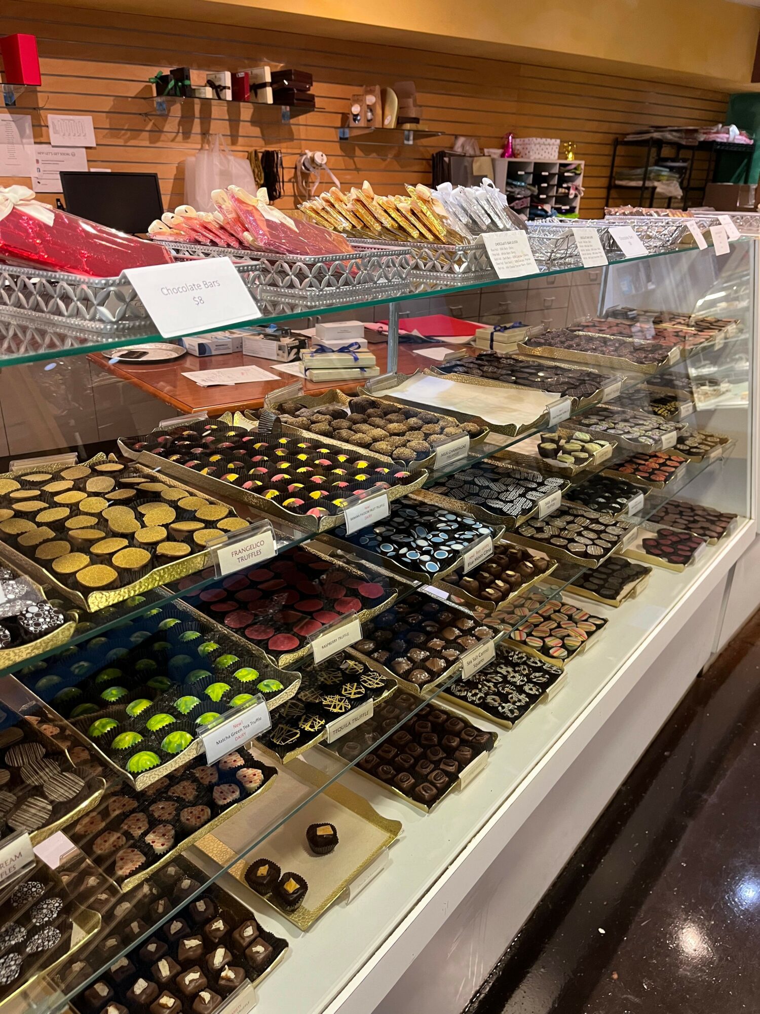 Display of assorted chocolates and confections in a store, arranged in rows on a glass counter with shelves in the background.