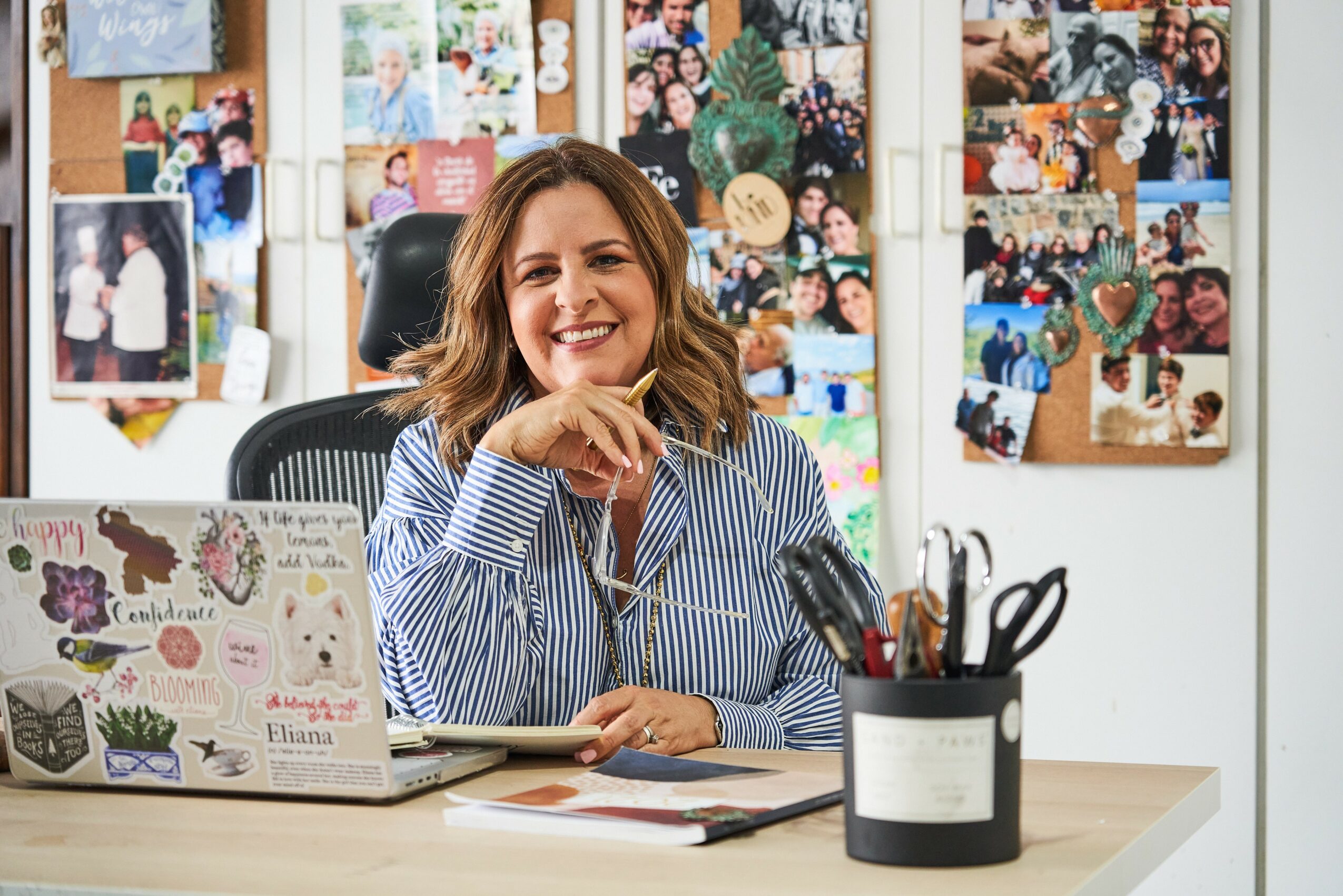 Woman smiling at desk with laptop and stationery, behind a collage of photos on the wall.