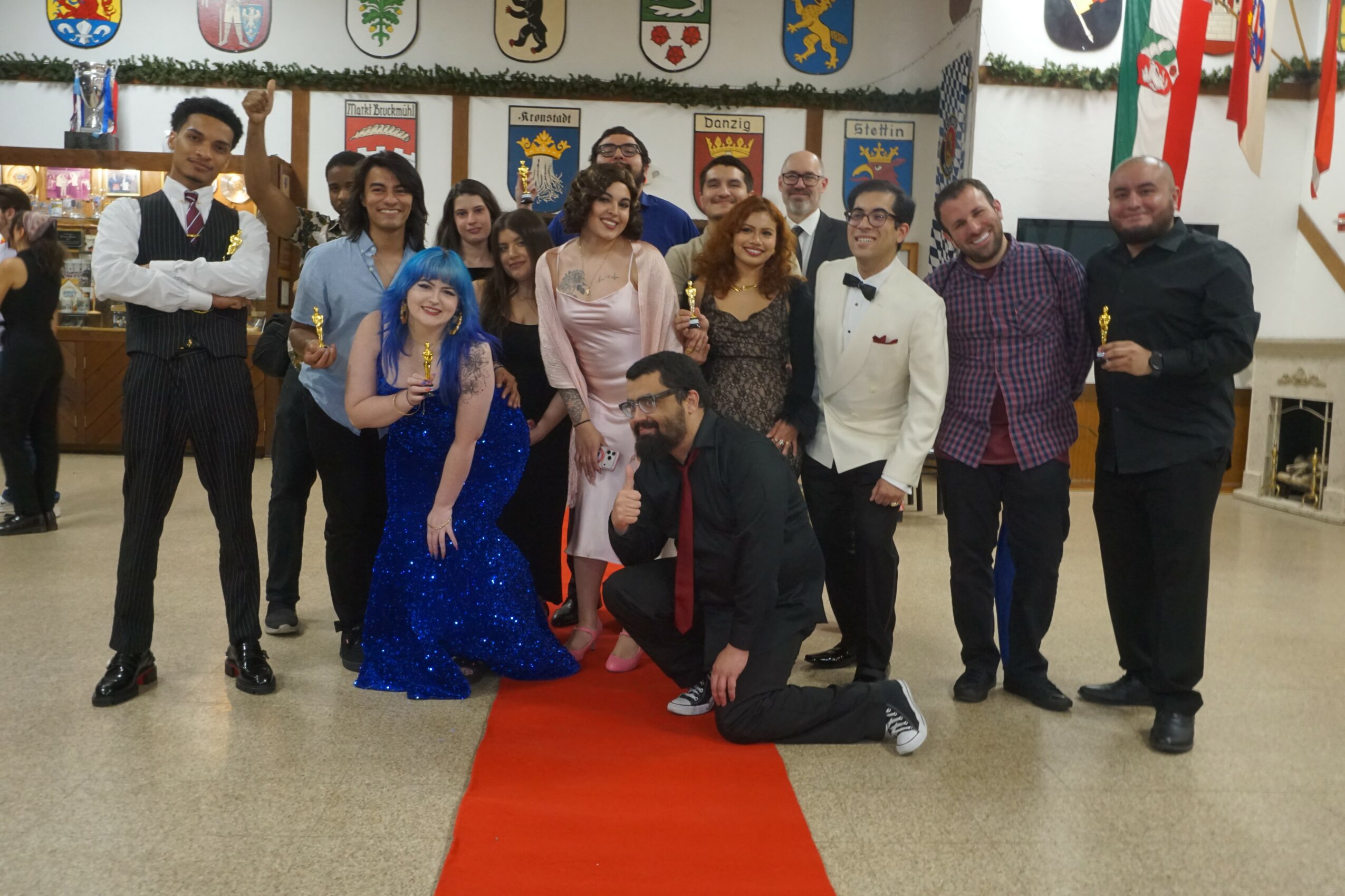 Group of people in formal attire posing indoors with banners and decorations, some standing and one kneeling on a red carpet.