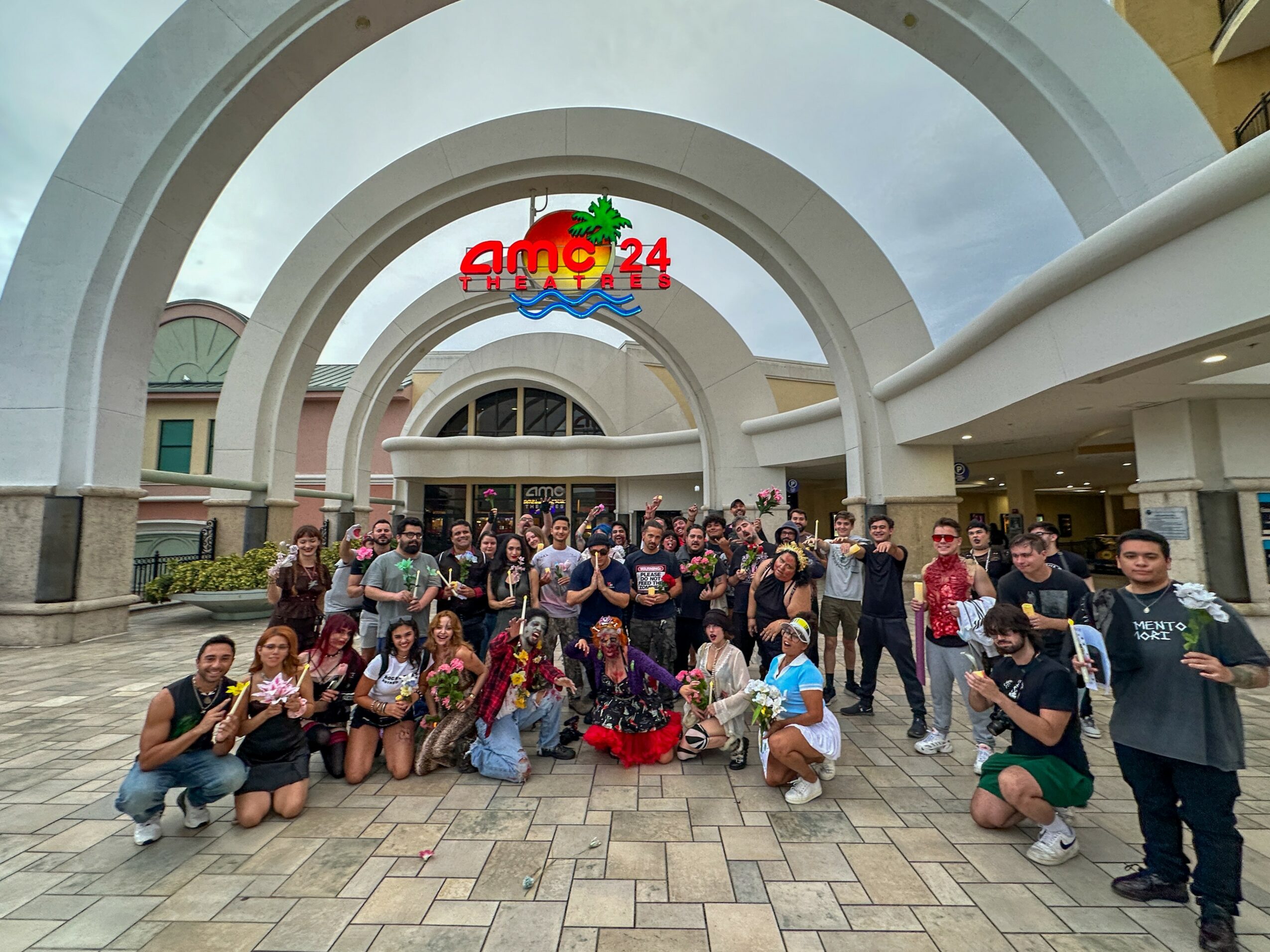Group of people gathered outside a building with large white arches and a colorful sign overhead.