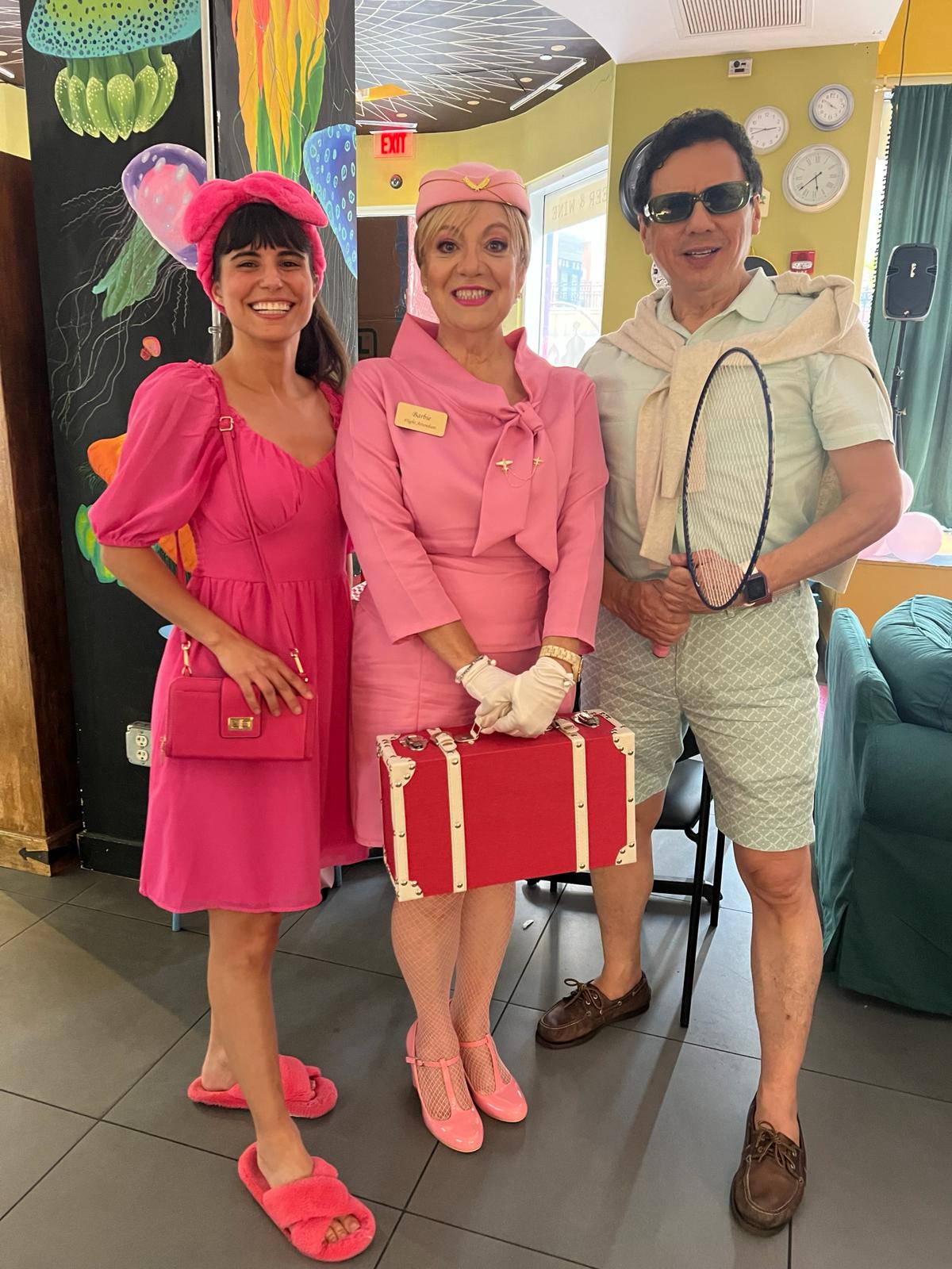 Three people dressed in vintage pink costumes, smiling indoors with colorful decorations and a green wall in the background.