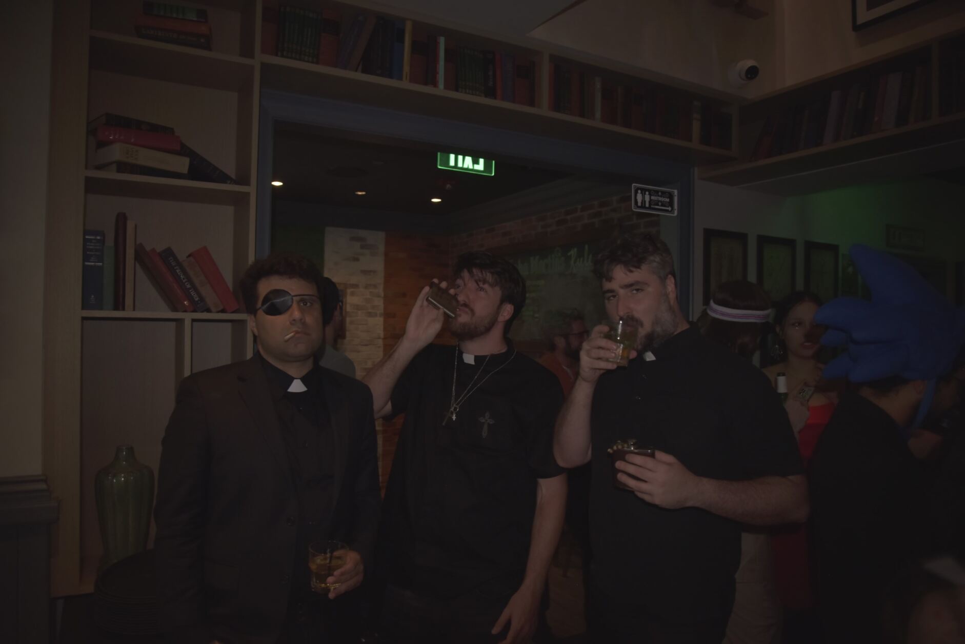 Three men standing in a room, two drinking from glasses, one wearing sunglasses, with bookshelves and a green exit sign behind them.