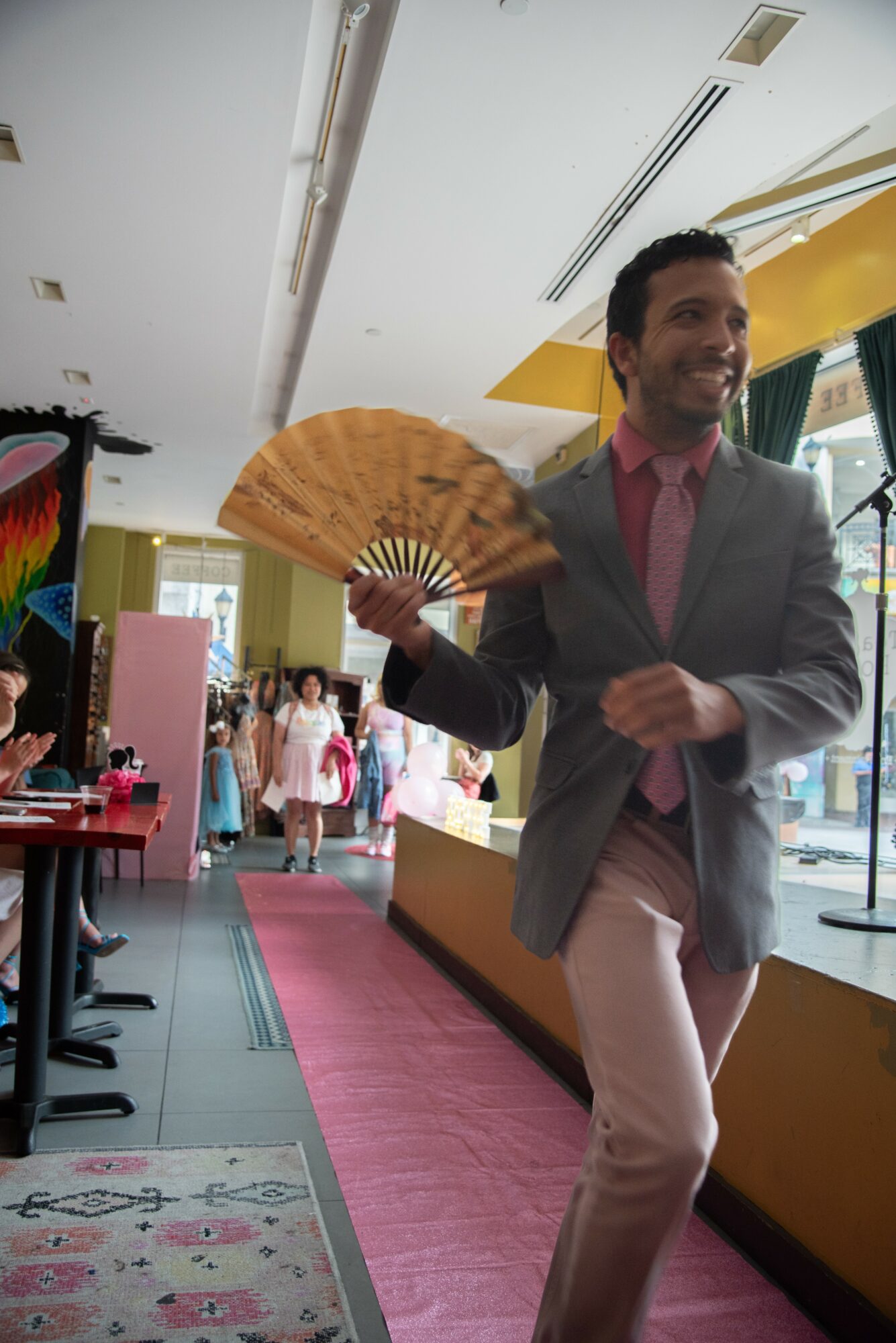 Man in gray blazer and pink shirt holding a fan, smiling, inside a colorful indoor space.