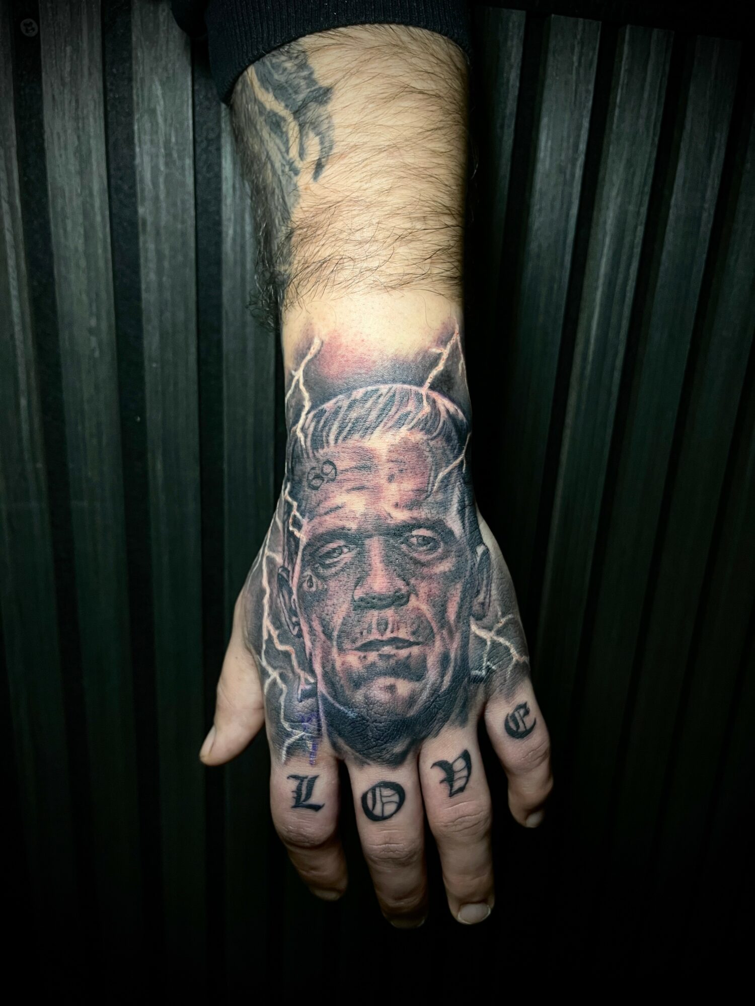 Tattooed hand with a portrait of a man and symbols on fingers, against a dark background.