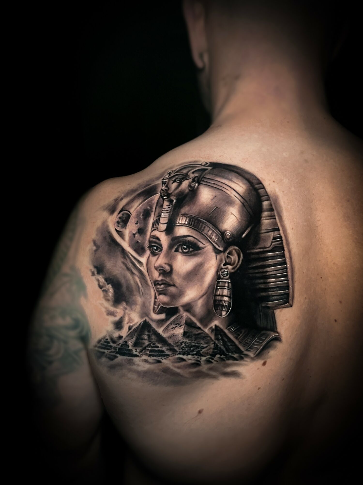 Tattoo of an ancient Egyptian woman with a headdress on a person's shoulder blade, with mountains and clouds in background.
