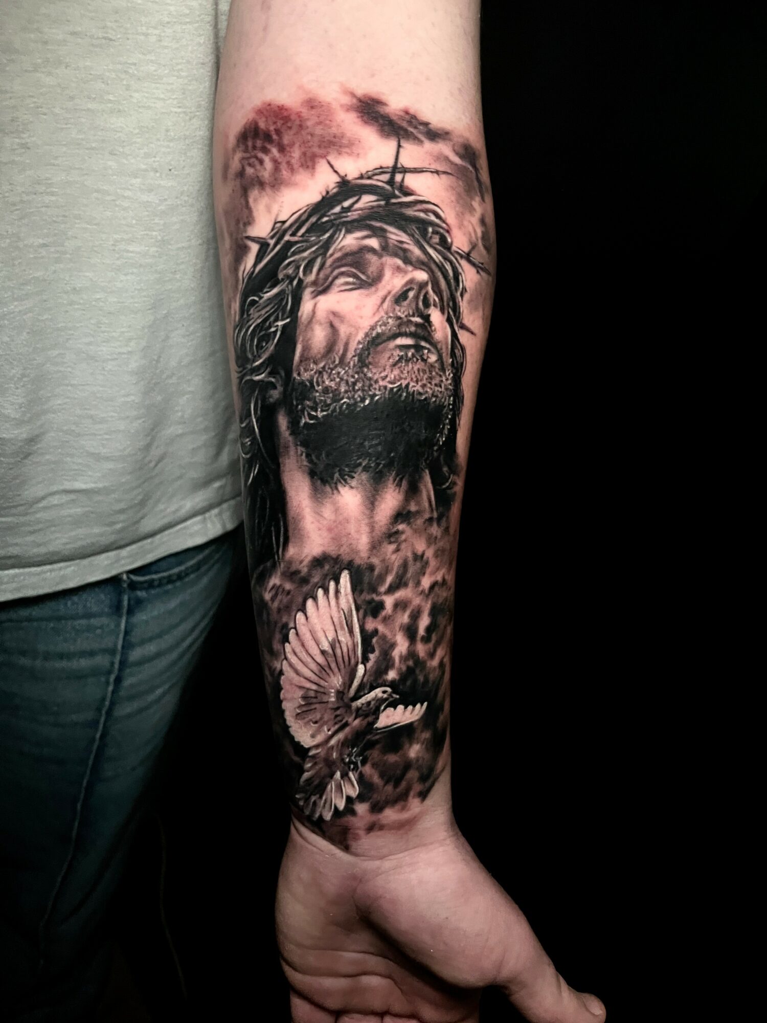 Tattoo of Jesus with crown of thorns, looking upward, with a dove below and cloudy sky background.
