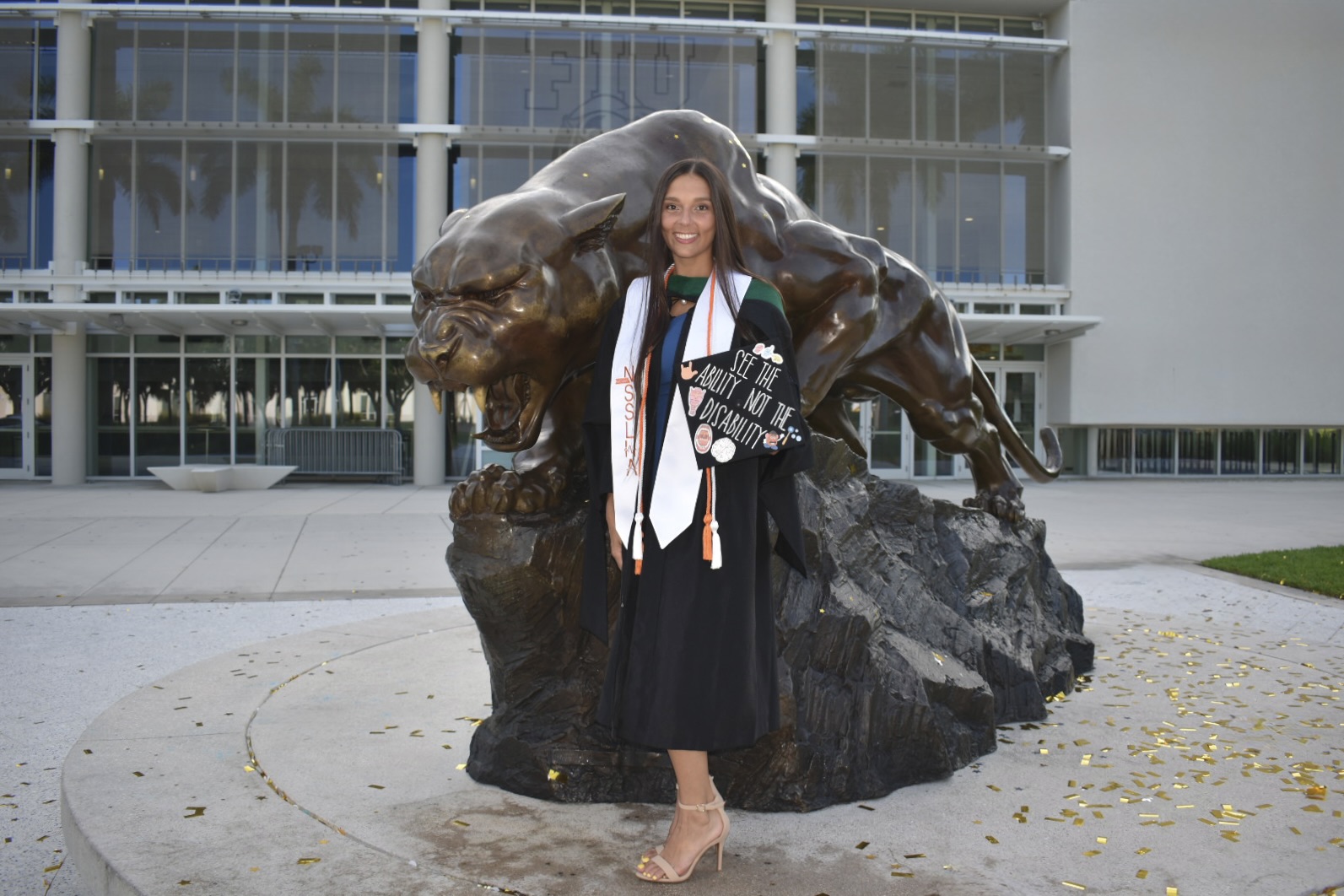 Woman in graduation gown and cap standing in front of a large panther sculpture outside a modern building.