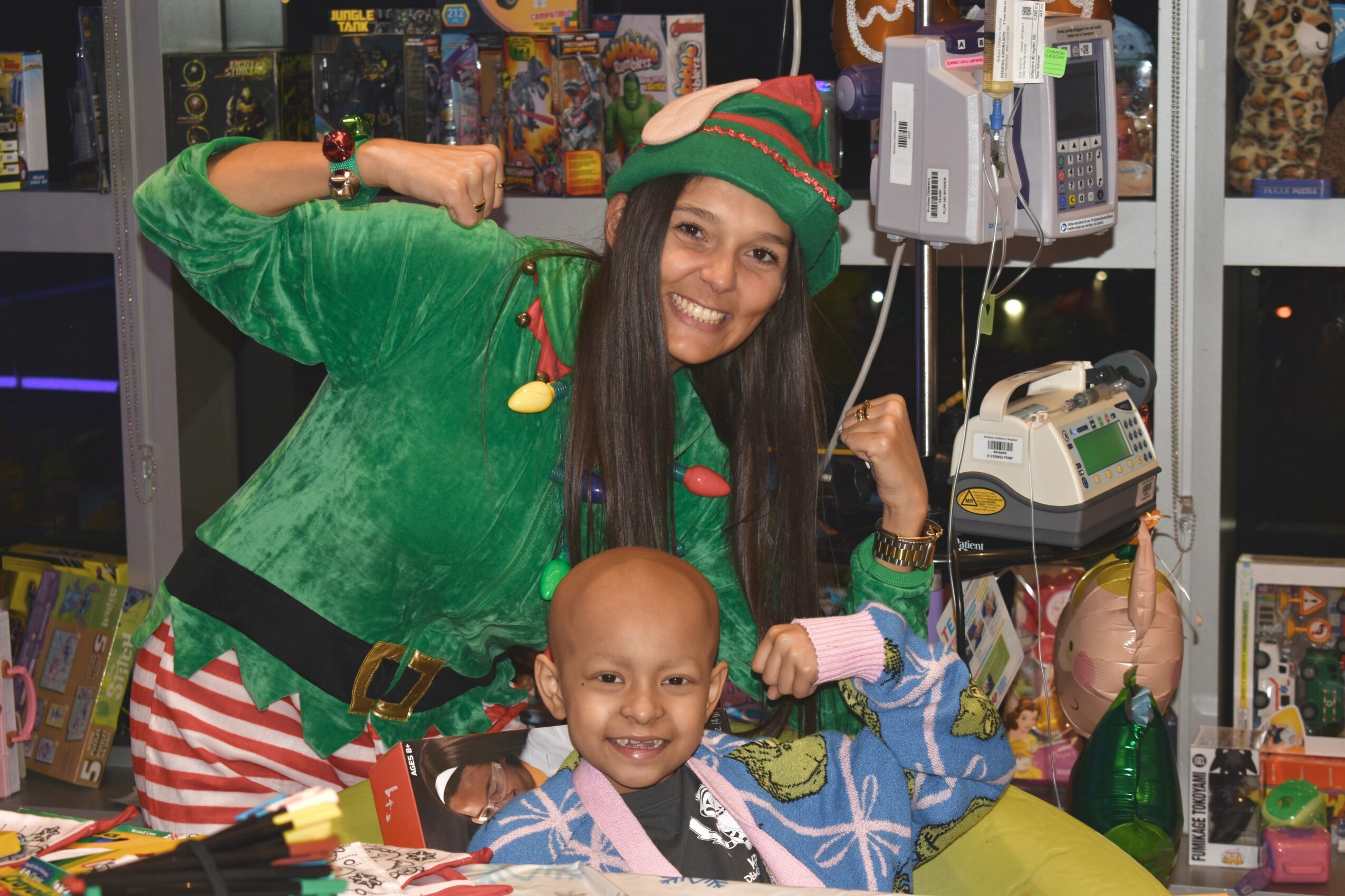 Woman dressed in a green holiday sweater and elf hat smiling with a young child in a festive setting.