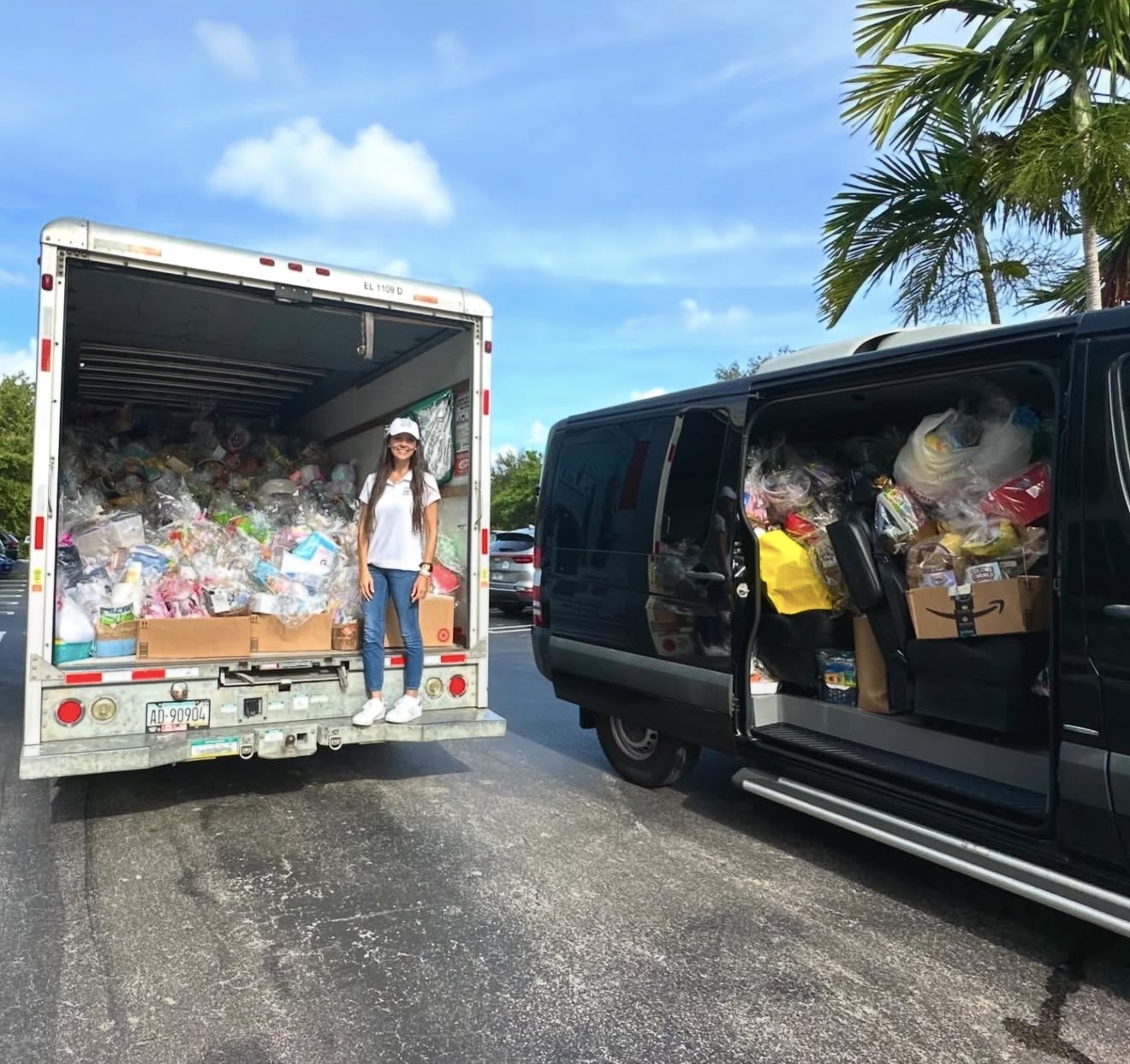 Person standing between two vehicles filled with bags of supplies, palm trees and blue sky in background.