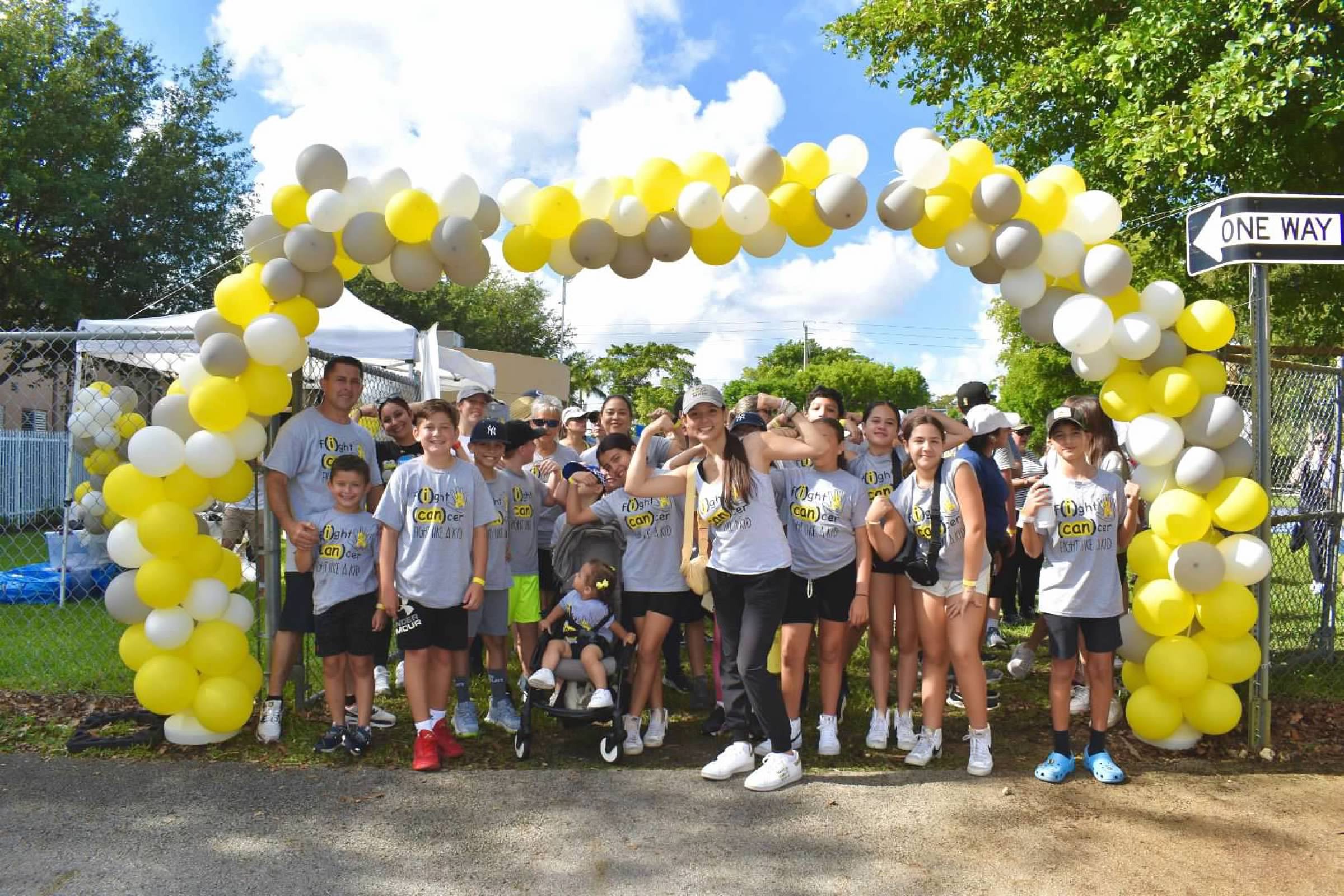 Group of children and adults gathered under yellow and white balloon arch outdoors during daytime.