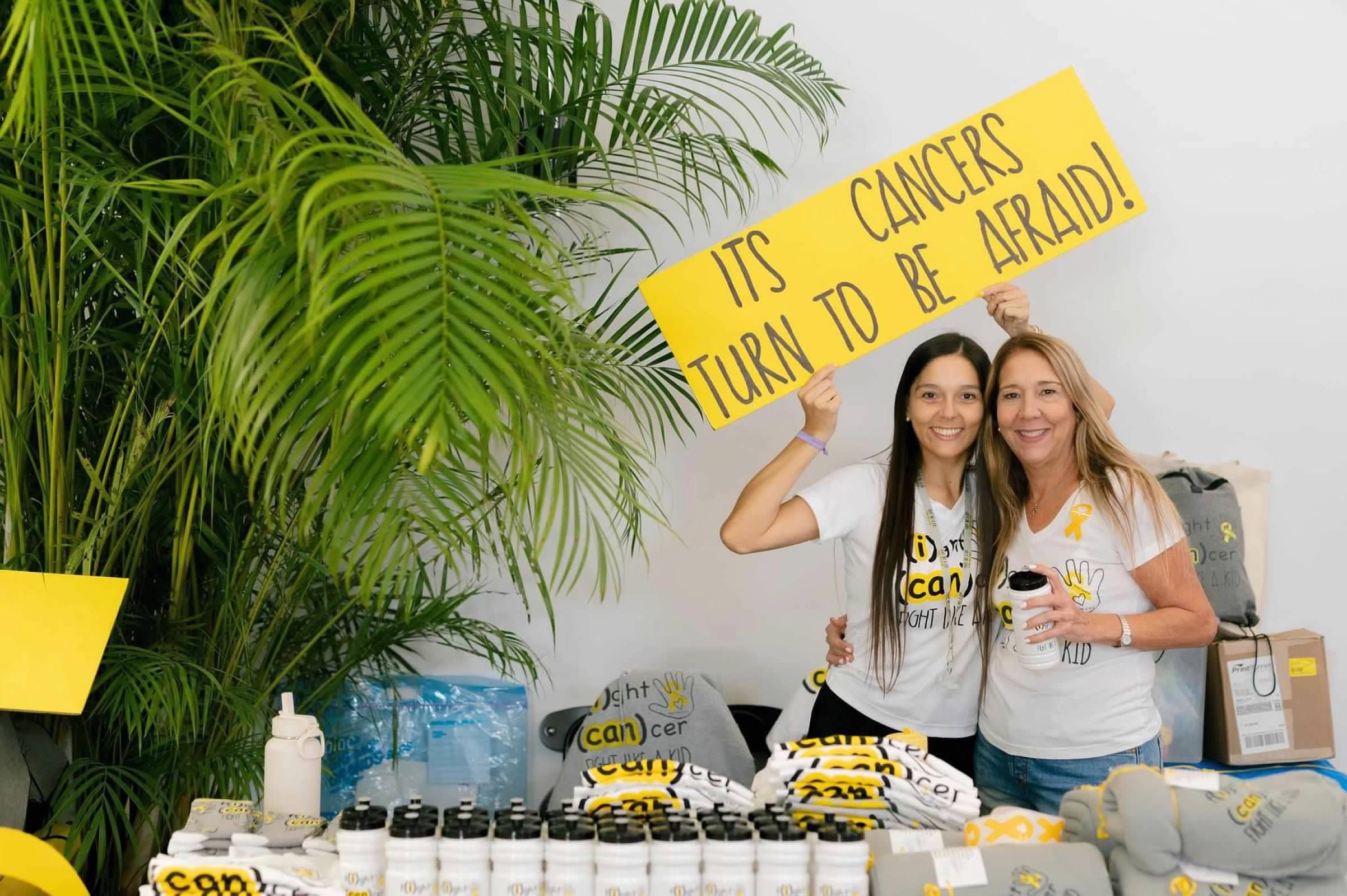 Two women smiling and holding a yellow sign that says 'Cancers turn to be afraid!' in front of a large green plant.