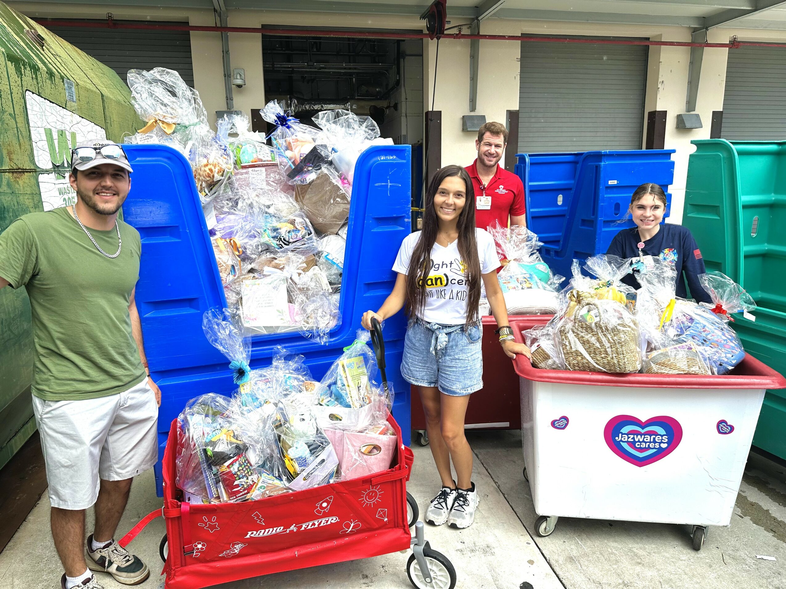 Four people stand in front of overflowing recycling bins filled with plastic and paper donations, outdoors in an industrial area.