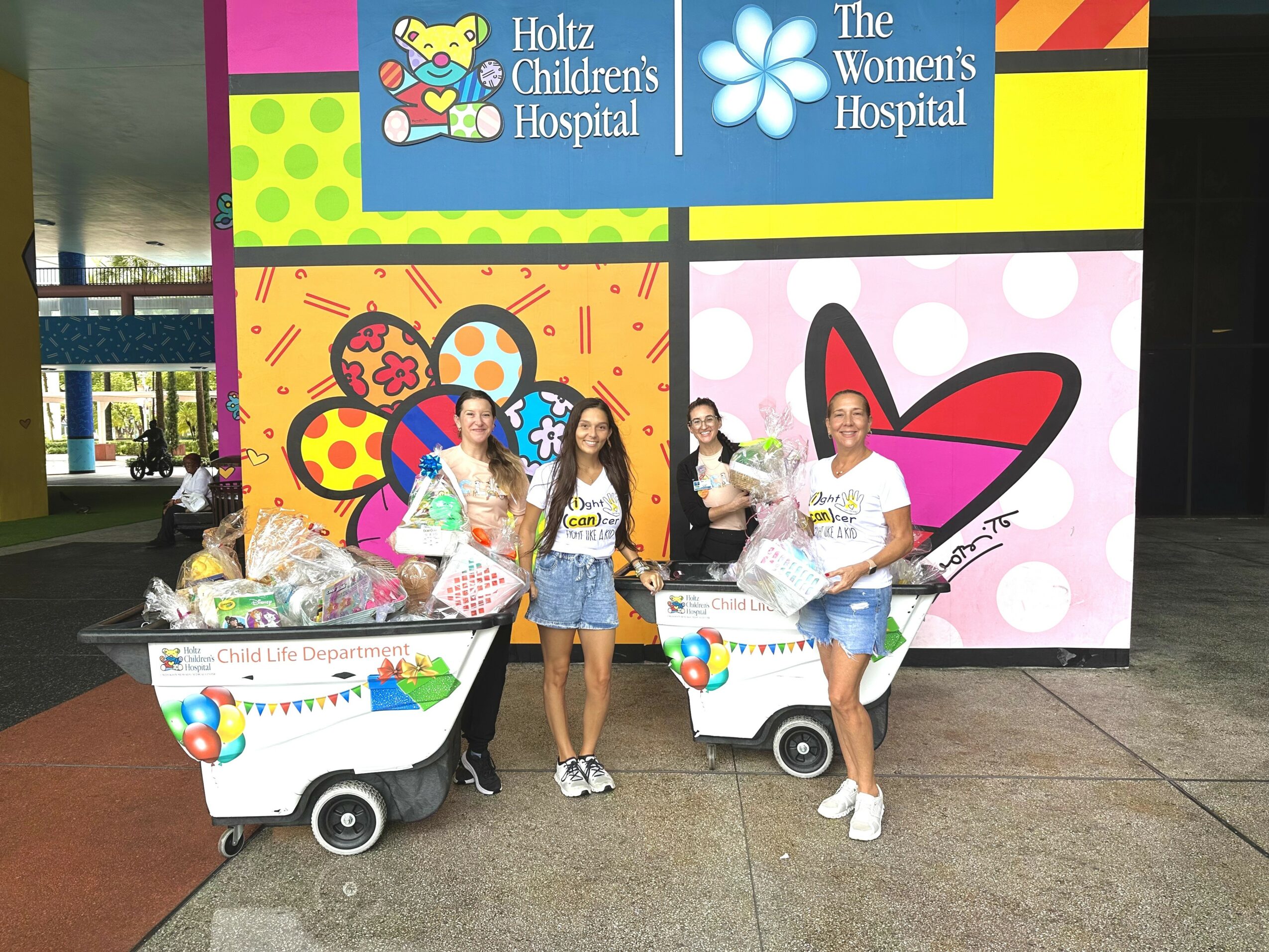 Four young women stand in front of colorful hospital signs, two with shopping carts filled with toys and gifts.
