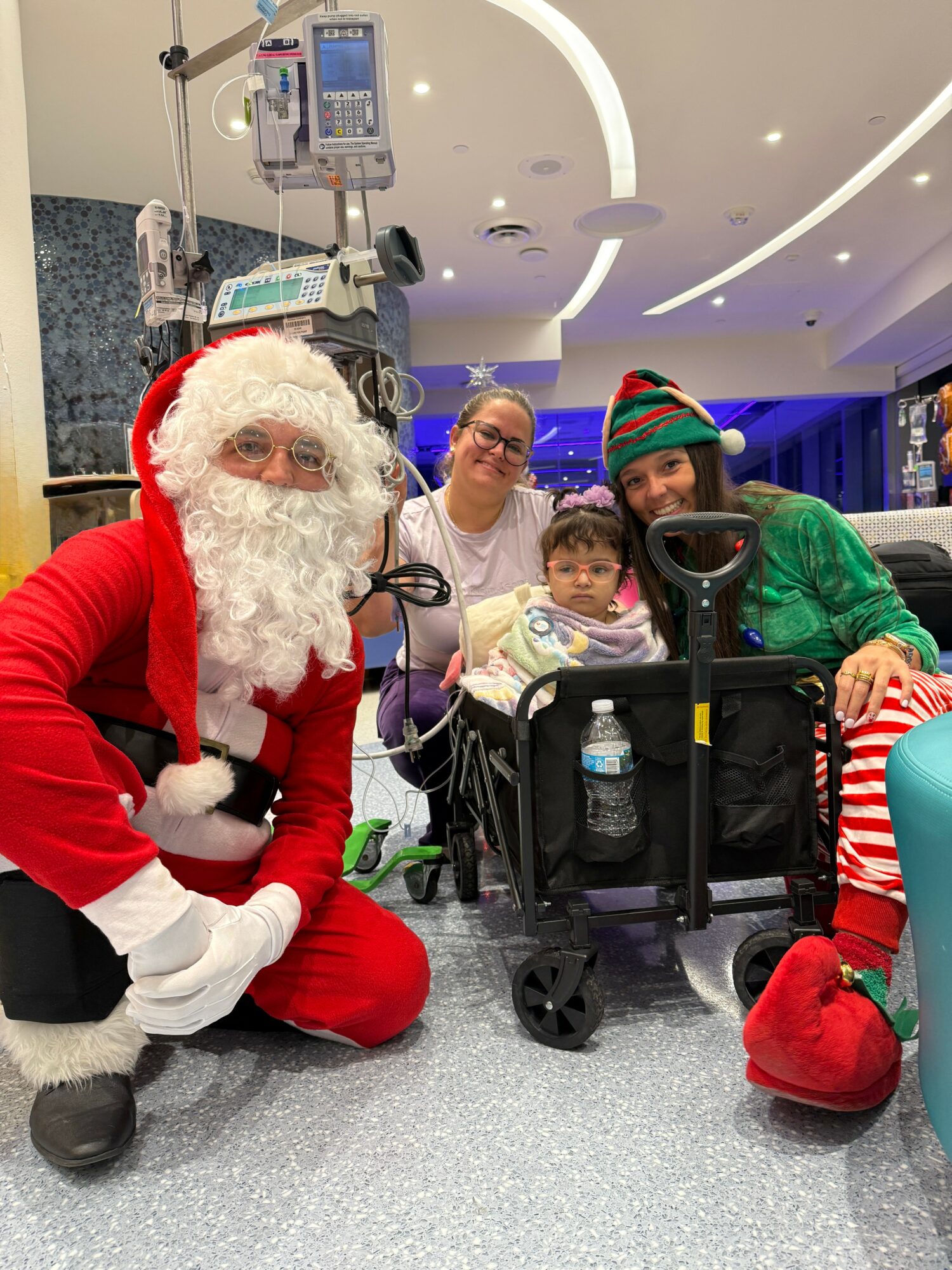 Person dressed as Santa Claus with children and caregiver in a hospital setting.
