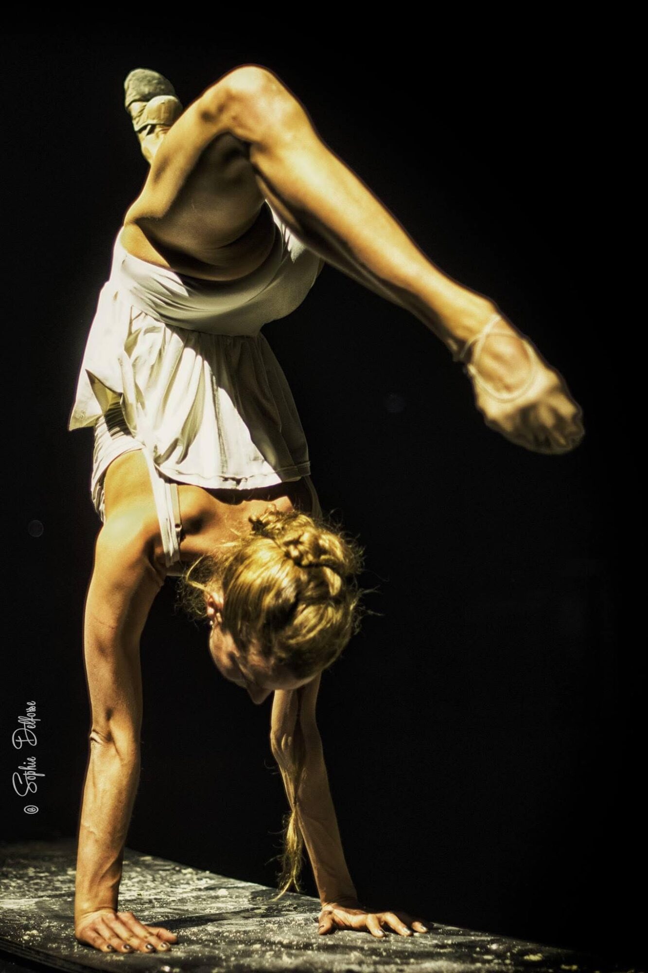 A person performing a handstand with legs bent and head near the ground, on a dark stage.