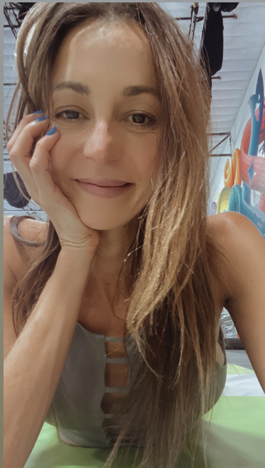 Woman with long hair resting her face on her hand, smiling softly, in a colorful indoor setting.