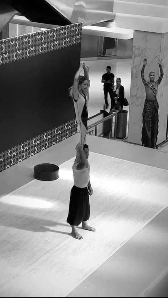 Two performers, one standing on the other's shoulders, raising arms in a dance or acrobatic pose, in a modern indoor space.