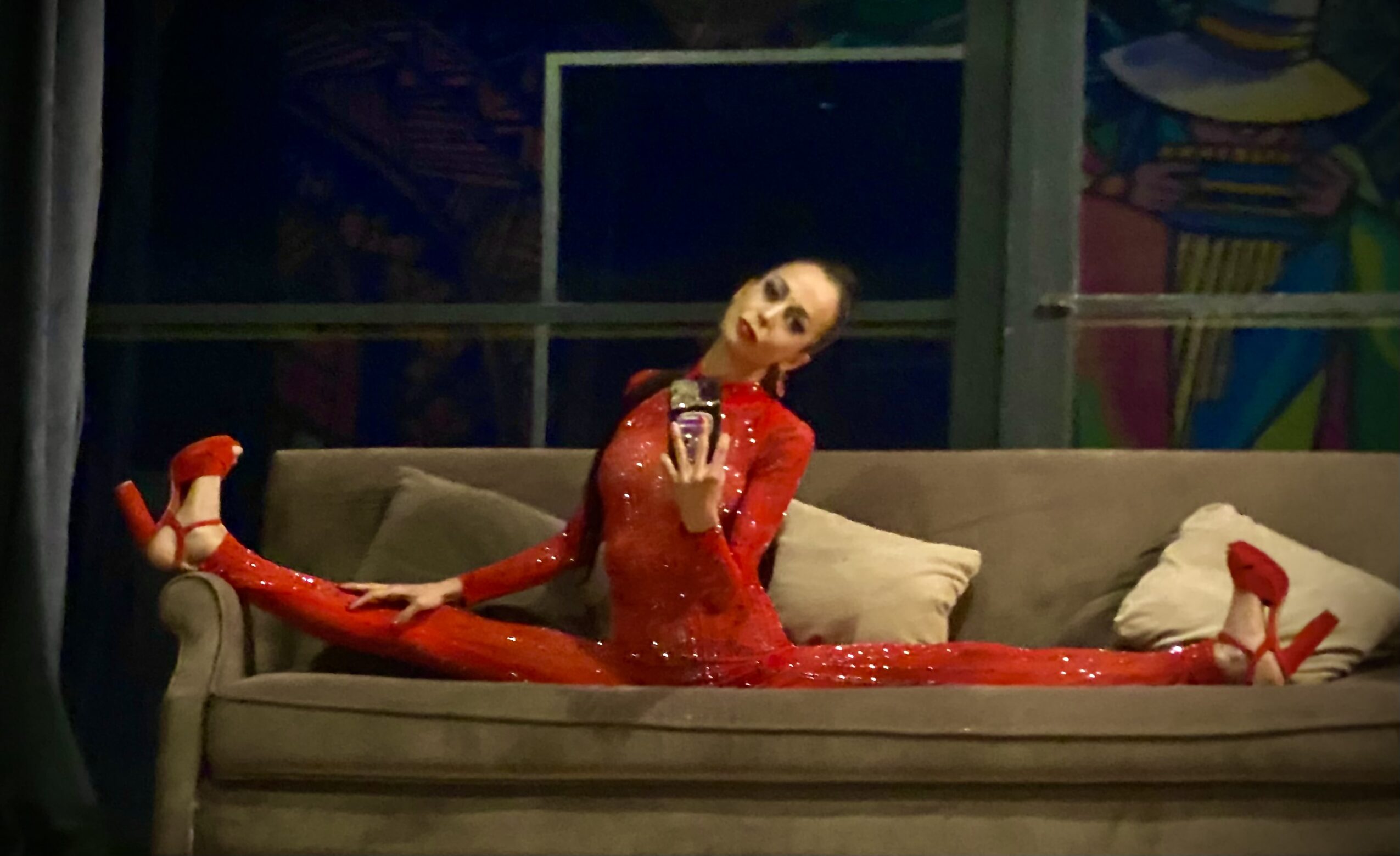 Person in red costume performing a split on a sofa with a backdrop of colorful artwork.