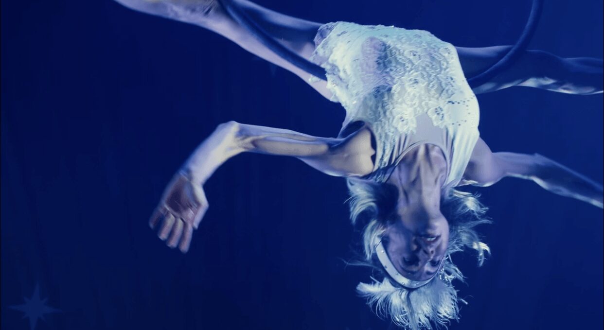 Person floating underwater with long hair and a flowing dress, arms extended, in a dark blue environment.