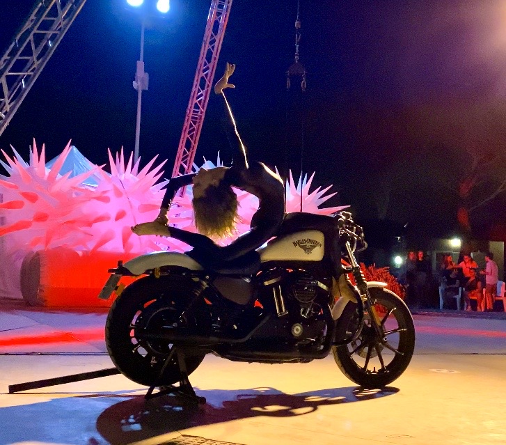 Person in costume with wings sitting on a motorcycle at night, illuminated by colorful lights.