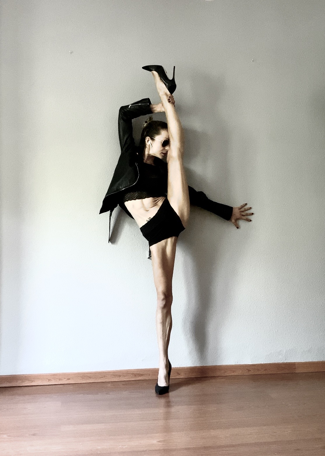 Person performing a high leg stretch against a plain wall, wearing black clothing and high heels.