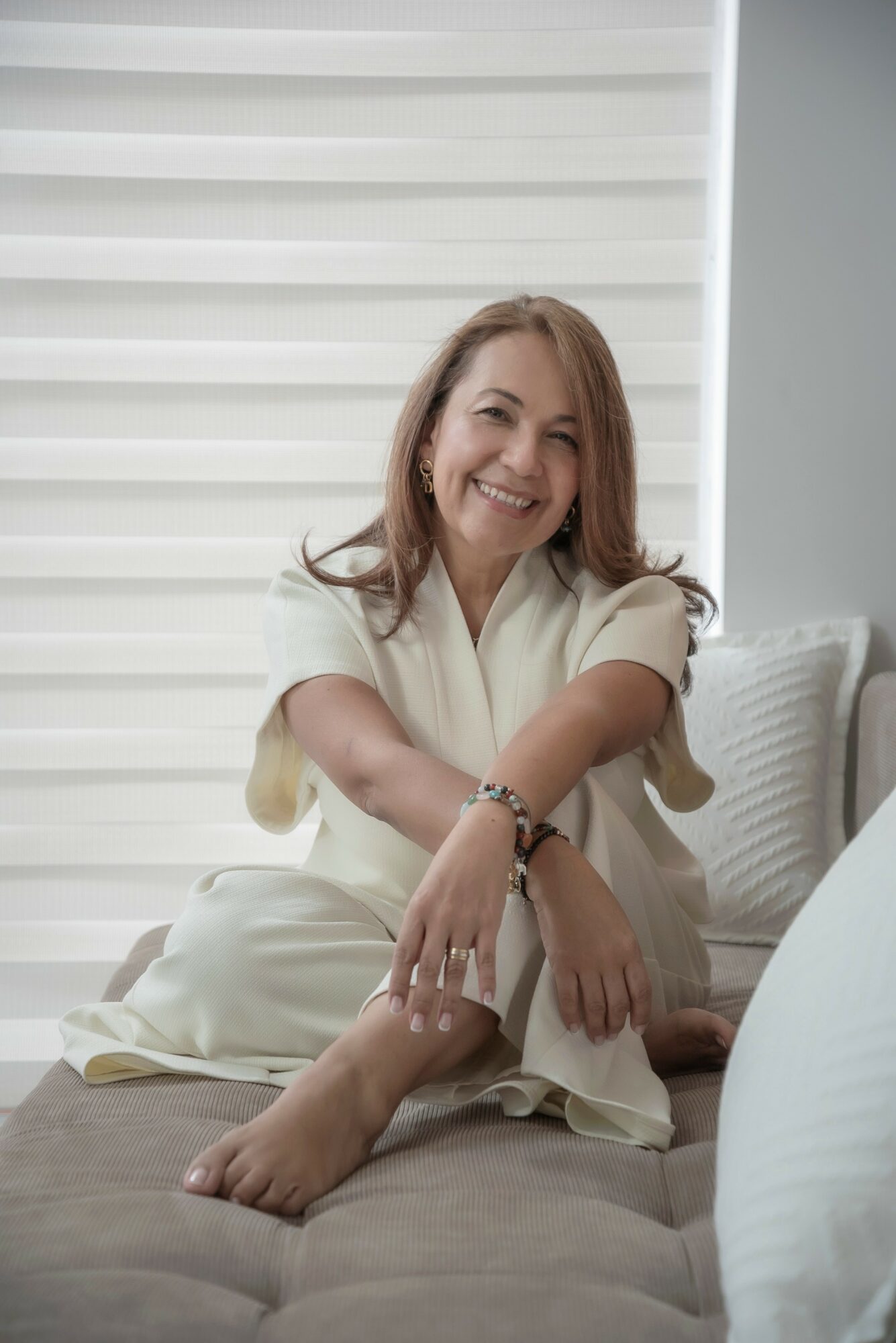Woman sitting on a bed, smiling, wearing a light-colored outfit, with a white background and window blinds behind her.