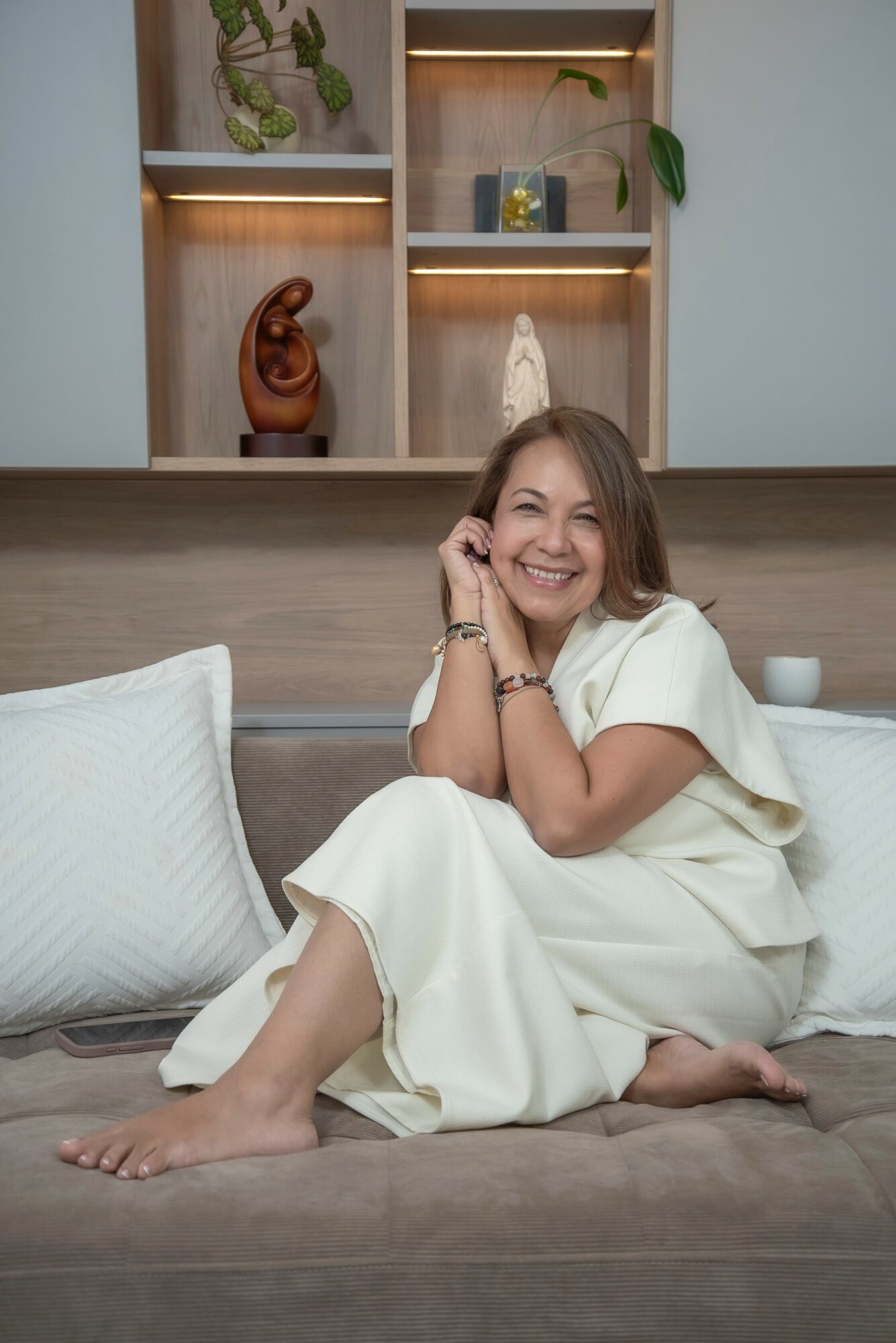 Woman sitting on bed, smiling, with shelves and plants in background.