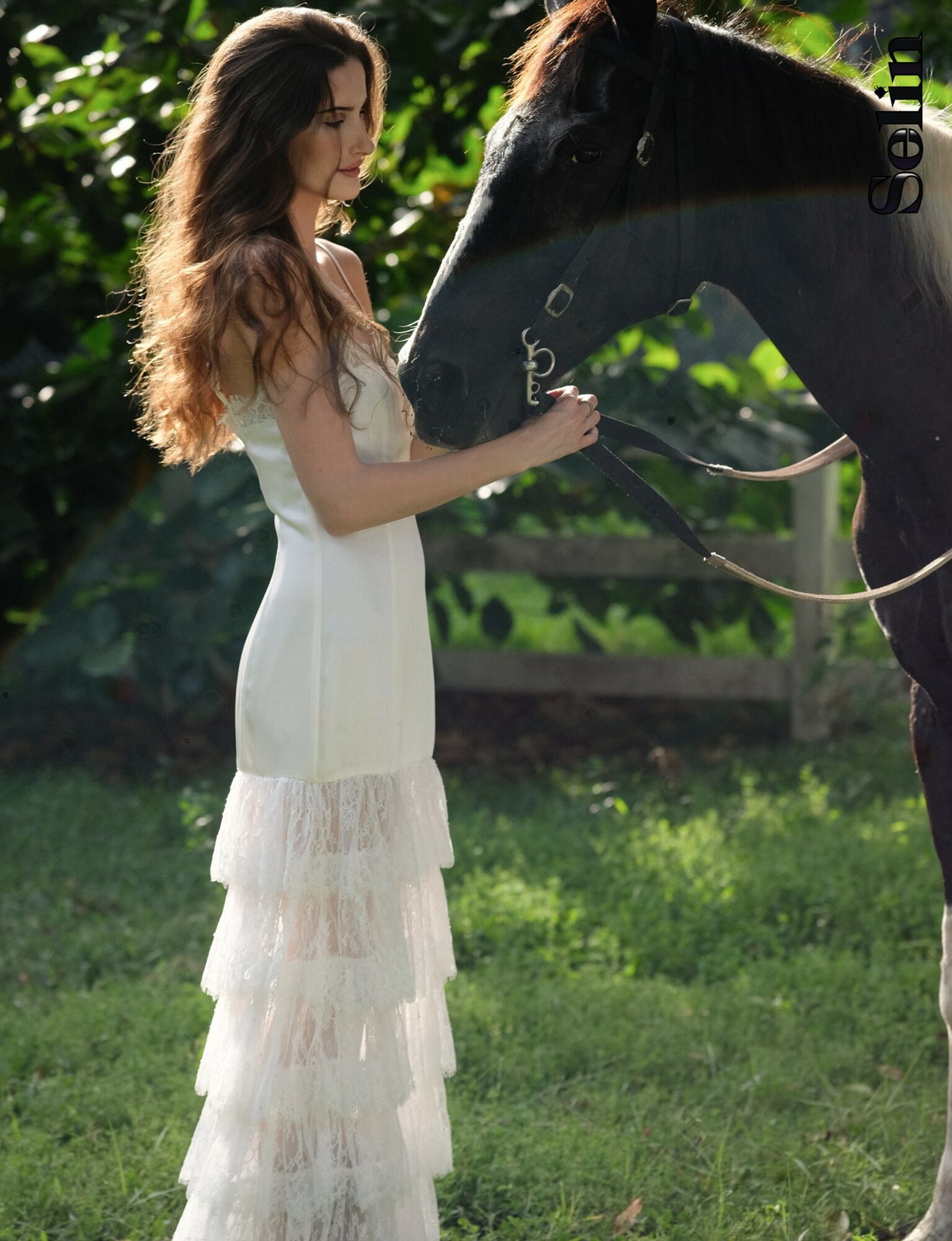 Woman in white dress holding a black horse's reins outdoors with green foliage background.