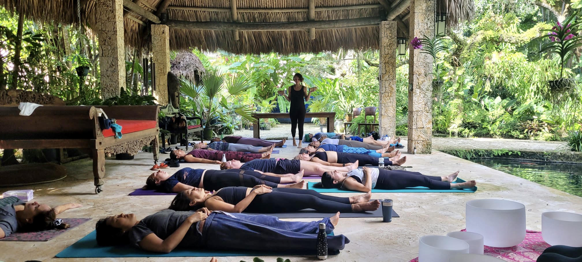 Group of people practicing yoga outdoors under a thatched roof, lying on mats with a instructor standing nearby.