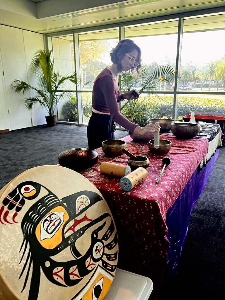 Person preparing food or craft at a table with bowls and tools, in a room with large windows and plants.