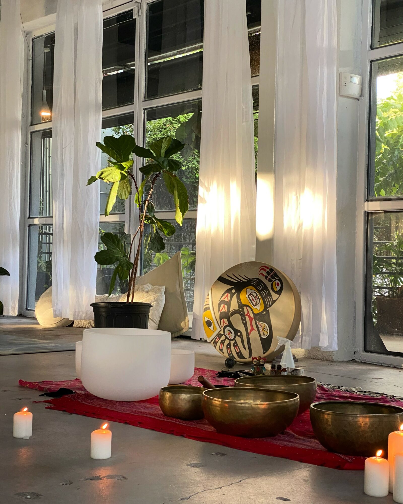 Indoor scene with a potted plant, singing bowls, candles, and sheer curtains by windows with greenery outside.