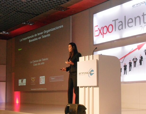 Woman standing on stage near a podium, speaking at a conference with a large screen behind her displaying text.