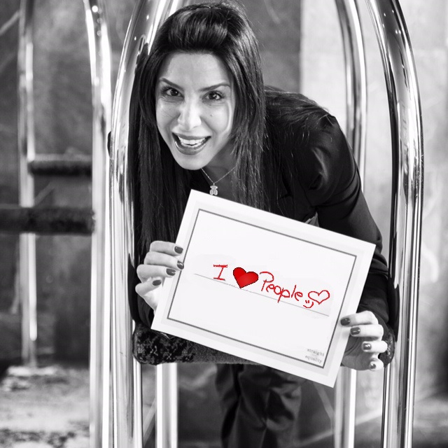 Woman with long hair smiling, holding a sign that says 'I love People' with a heart symbol.
