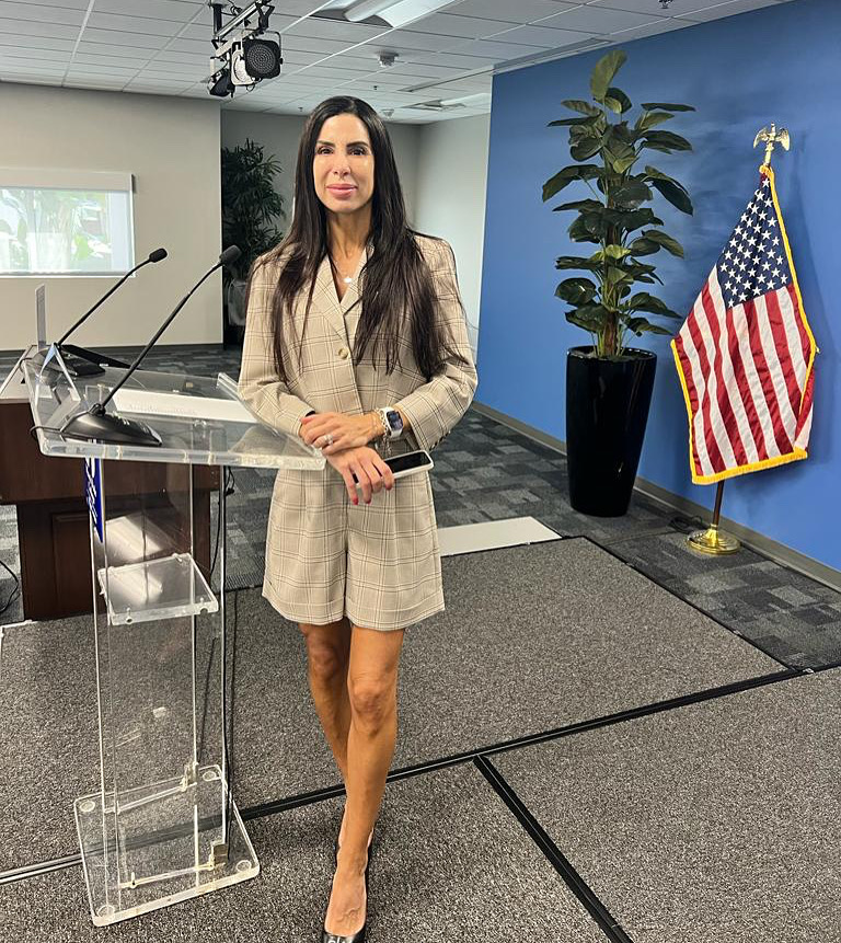 Woman in beige blazer dress standing near a clear podium with a microphone, American flag, and potted plant behind her.