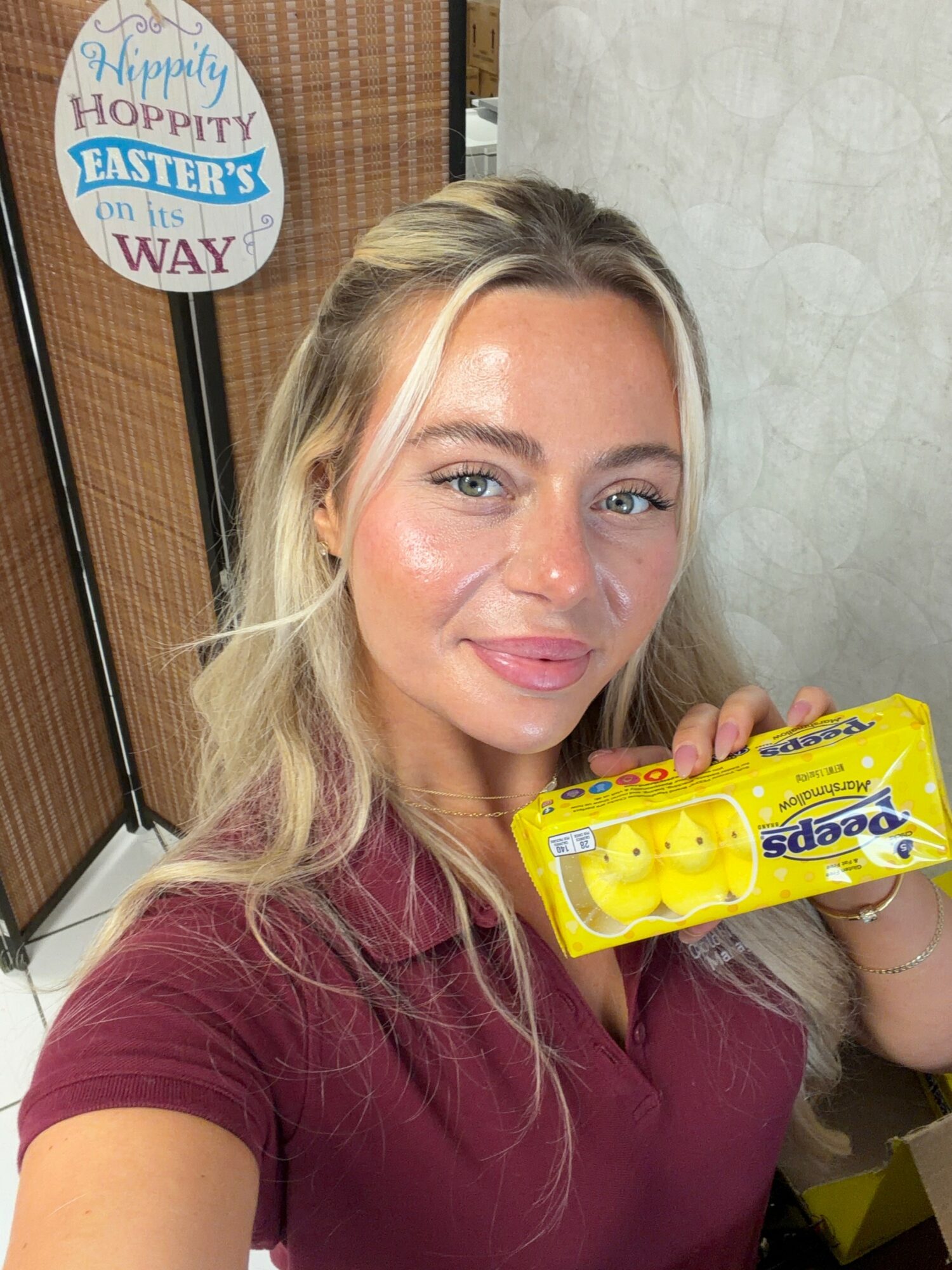 Young woman with blonde hair holding a yellow pack of eggs, smiling, in front of a wall with Easter decoration.